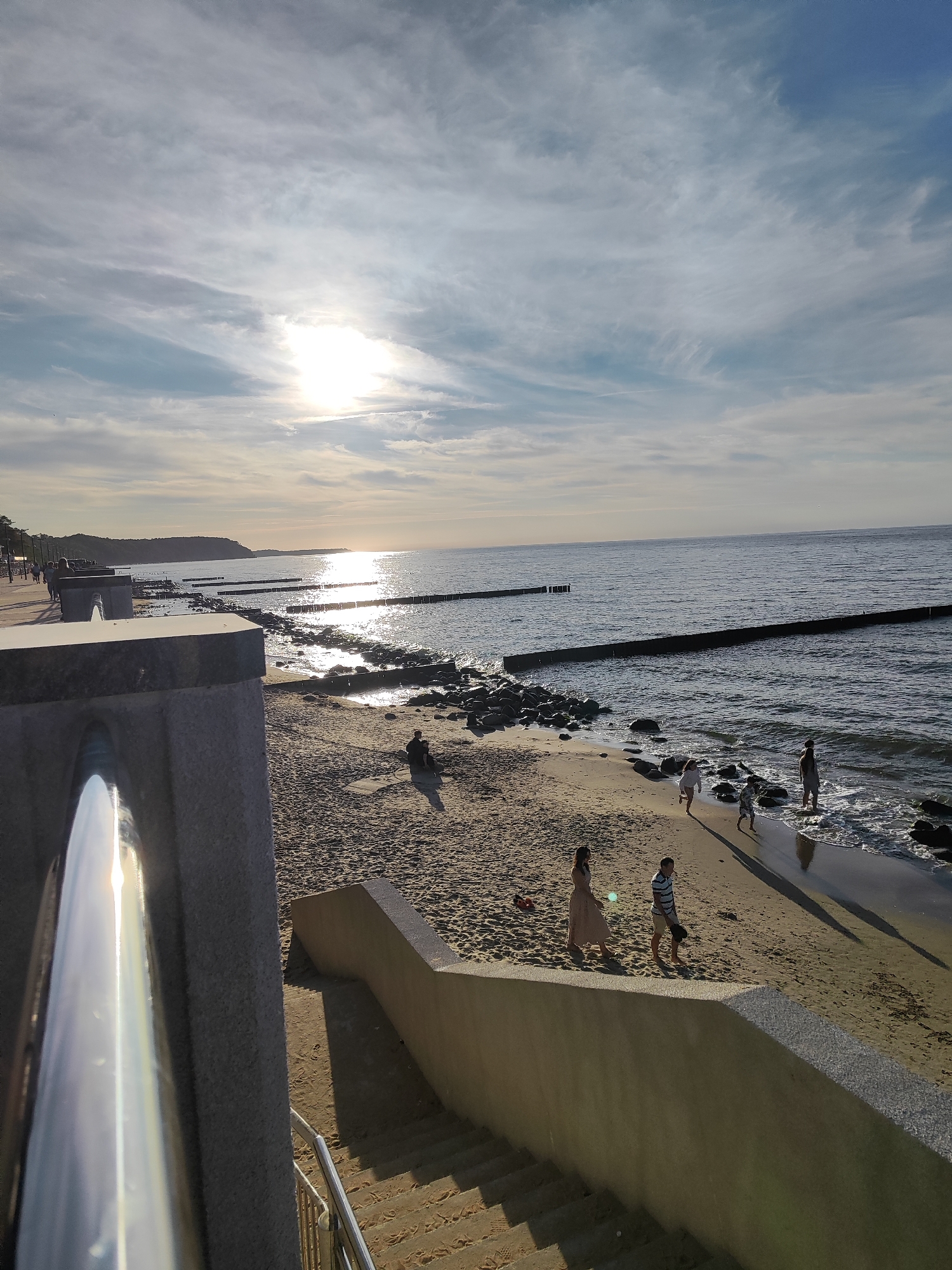 The image depicts a sunlit beach in Svetlogorsk, Russia, with a wide expanse of the Baltic Sea. The sun, partially obscured by thin clouds, casts a bright reflection on the water. Several wooden breakwaters are visible in the sea, stretching from the shore into the distance. The sandy beach is populated by a few individuals and small groups. In the foreground, a concrete walkway with a metallic railing overlooks the beach and sea. A set of concrete stairs leads down to the sand from the walkway. The sky is partly cloudy with streaks of cirrus clouds. The overall atmosphere is one of a calm, late afternoon at a coastal resort.