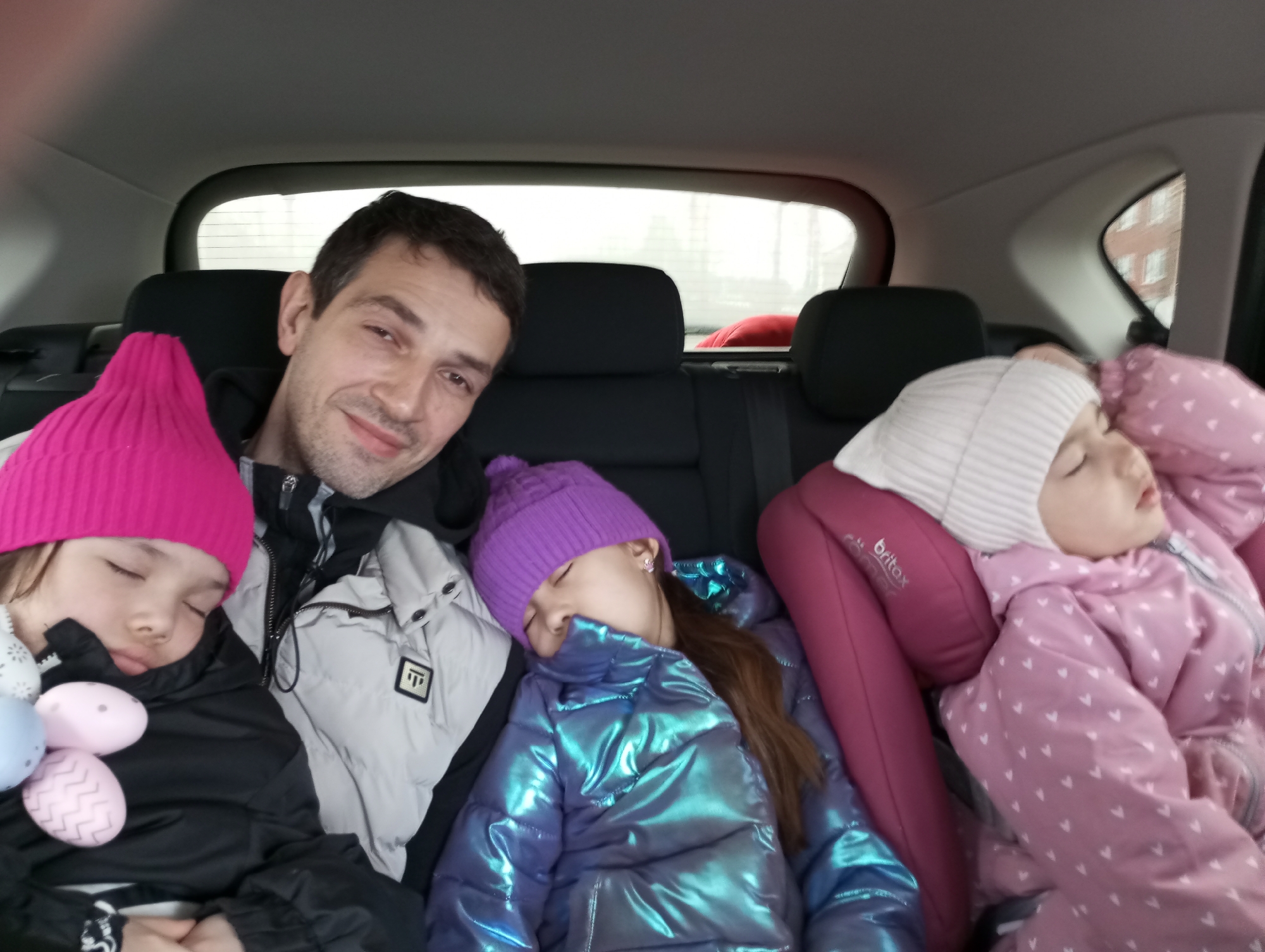 A medium, indoor shot depicts a man and three young children in the back seat of a car. The man is fair-skinned with short dark hair and stubble. He is wearing a light grey and black jacket. He is looking towards the camera with a slight smile. The child to his left wears a bright pink knit hat and a black jacket, and is holding several painted eggs. The child next to him wears a shiny blue-purple jacket and matching purple knit hat. The child on the right is wearing a patterned pink jacket and a white knit hat, and is secured in a Britax car seat. All three children are sleeping. The car interior is grey, with the rear window showing a blurred view of an exterior scene that appears to be in an urban environment, potentially near Фоки, Russia.