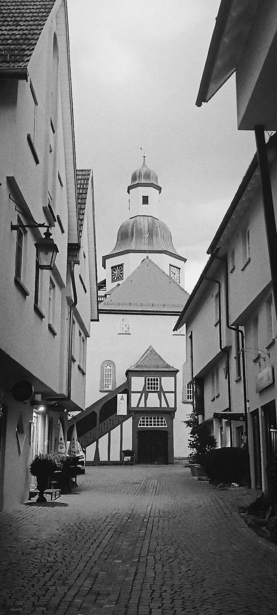 The black and white, vertical, medium-angle shot captures a narrow cobblestone street in Winnenden, Germany, leading towards a church with a prominent dome and clock tower. The sky is overcast. On the left, a multi-story building features windows and a decorative lantern. On the right, another building with a dark gutter runs parallel to the street. The street itself is paved with uneven cobblestones. In the foreground, a stone planter filled with greenery and some outdoor seating are visible. The central focus is the church, characterized by its light-colored facade, a pitched roof leading up to a tiered tower topped by a dome, and two visible clock faces. A covered staircase with wooden railings ascends to an entrance on the church's lower level, which is adorned with half-timbered architectural elements and a gabled roof. The scene appears to be devoid of people, suggesting an early morning or late evening setting, or a quiet period during the day.