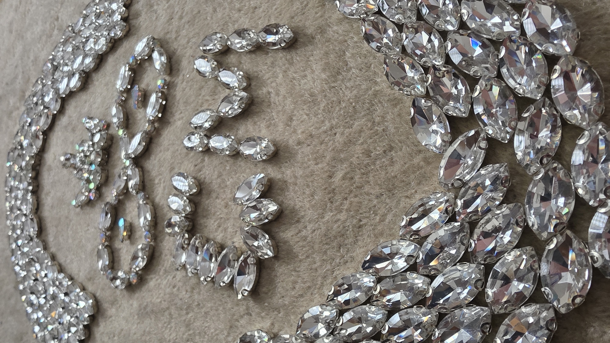 This close-up, high-angle shot captures a decorative arrangement of numerous clear, faceted rhinestones set against a soft, textured beige or light brown fabric, possibly felt. The majority of the rhinestones are marquise-cut, featuring bright, sparkling reflections and occasional iridescent rainbow glints from their facets. Many are encased in visible metallic settings.
On the left side of the frame, a dense, curved border or chain of these rhinestones runs along the edge, extending from the bottom left upwards. In the central area, several rhinestones are arranged to form the letters "BIZE". These letters are composed of both marquise and some smaller, round or oval shaped stones. To the right, another large, curving cluster of marquise-cut rhinestones appears, suggesting a continuation of a border or circular pattern. The lighting is bright, emphasizing the reflective qualities of the stones. No people or specific activity are depicted; the image focuses solely on the static, jeweled design.