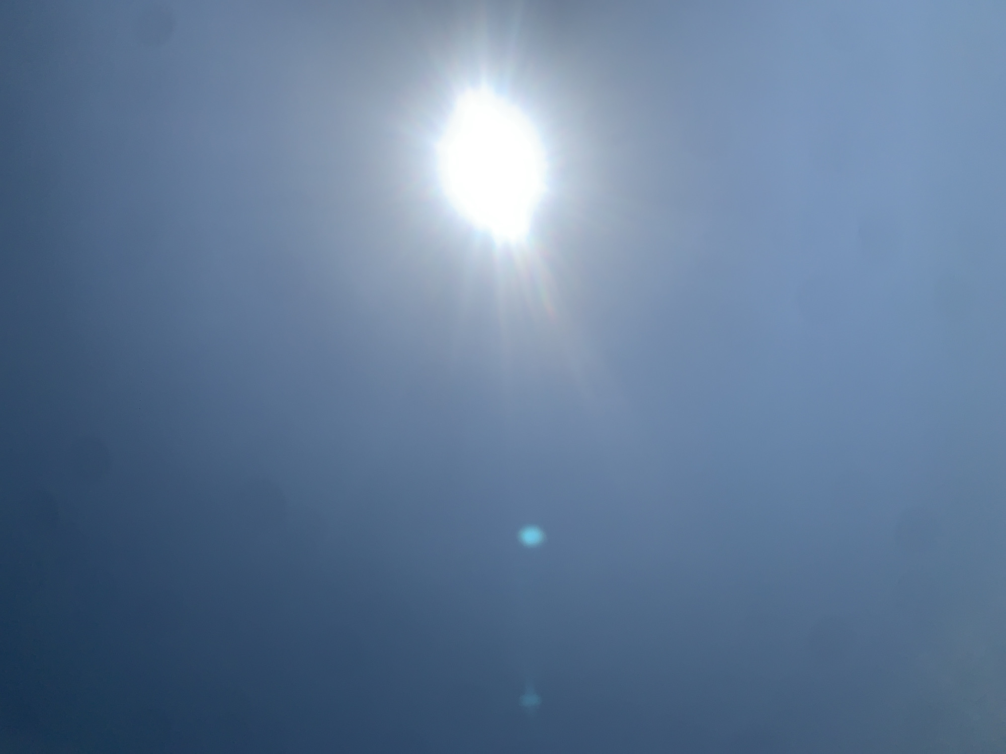 A bright sun shines in a clear blue sky, creating a lens flare that obscures much of the image. The sun is located in the upper right quadrant of the frame. Two faint, blue circular light flares are visible in the left quadrant of the image. The scene appears to be an outdoor environment, likely taken in Khazar Raion, Azerbaijan. No people or other objects are discernible in the image.