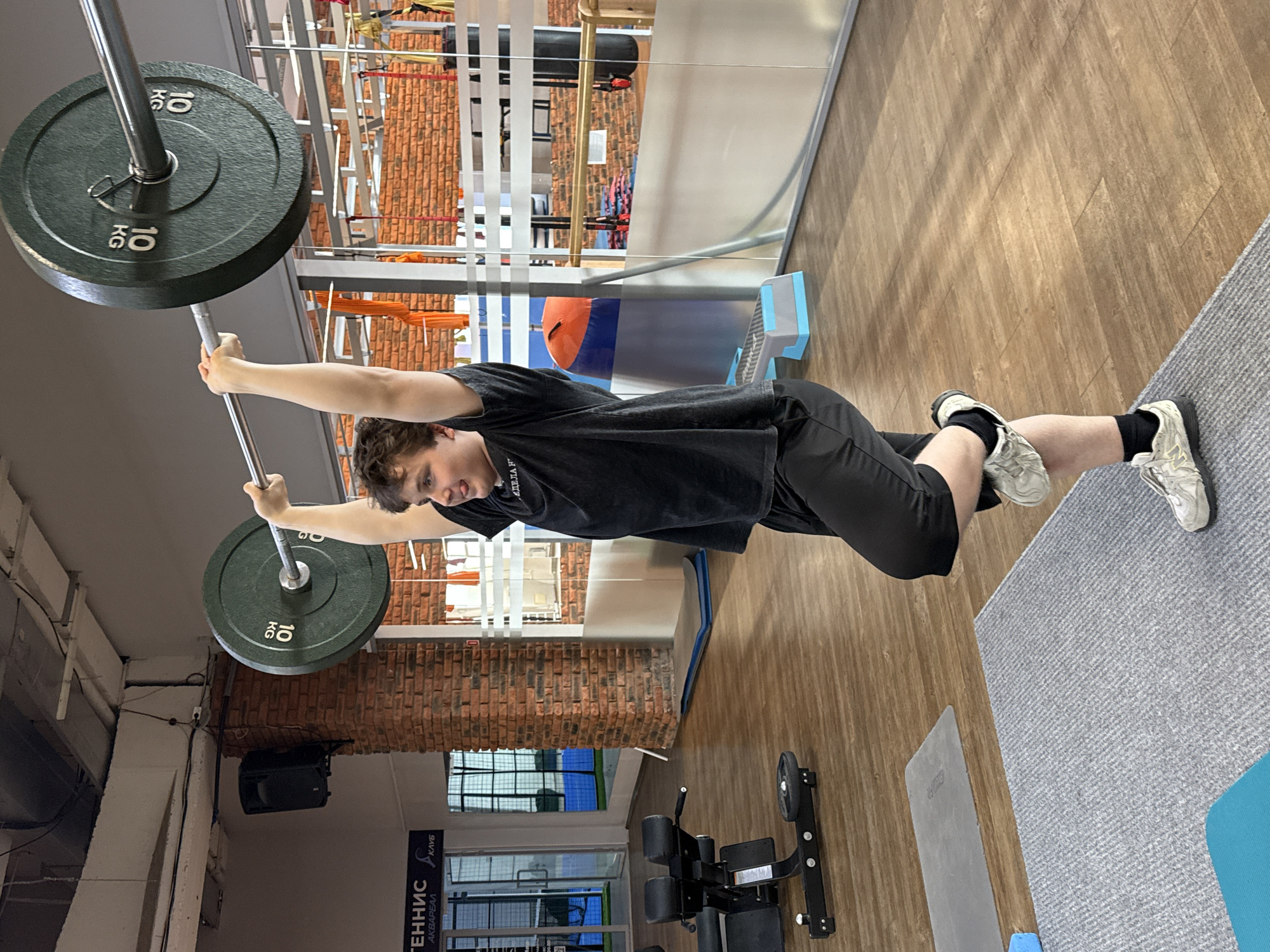 A young man is photographed in a gym. He is holding a barbell with 10 kg weights on each side above his head. He is wearing a black t-shirt, black shorts, and white sneakers with black socks. The gym has brick walls, windows, and various exercise equipment. The location is Chaikovsky, Russia, as evidenced by signage in the window.