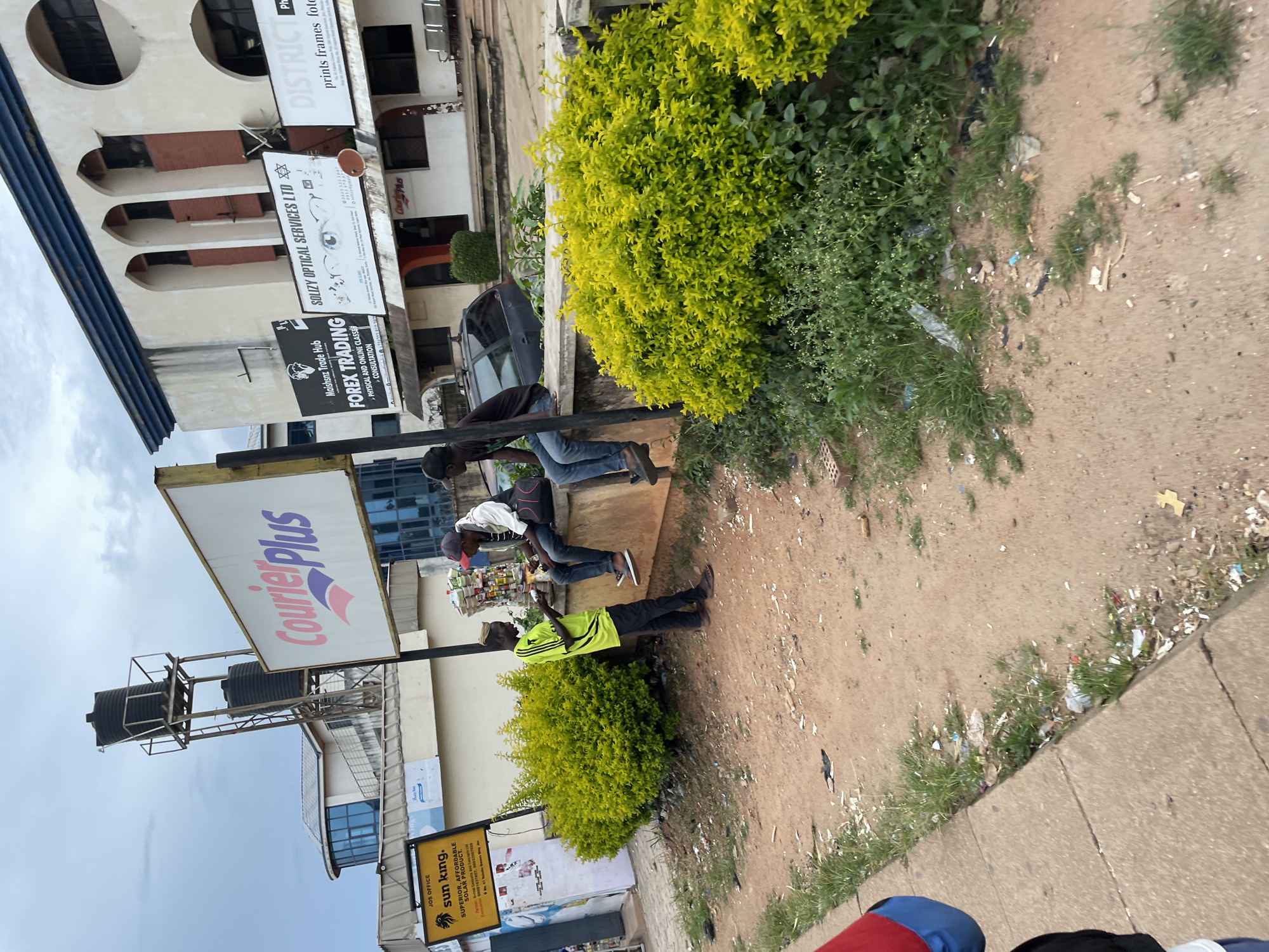 The image depicts a street scene in Jos, Nigeria, with three young men standing and sitting near a low concrete wall. A lush green bush borders the wall. One man, wearing a neon green shirt and black pants, stands facing a black car parked behind the wall. Another man, in a white jacket and dark pants, sits on the wall, looking down. A third man, also in dark pants, sits to his right on the wall, facing away from the camera.
Behind them, various signs are visible. A large white sign with "Courier Plus" in pink and purple letters is prominent. To the left, a yellow sign for "sun king." advertising solar products. Further to the right, a black sign reads "Maisknz Trade Hub FOREX TRADING." and a white sign for "SOLIZY OPTICAL SERVICES LTD." Another sign on the far right displays "DISTRICT prints frames foto".
A tall metal structure with two large black water tanks sits atop a building in the background. The ground is a mix of reddish-brown soil and scattered debris, including plastic waste and papers. The sky is overcast.