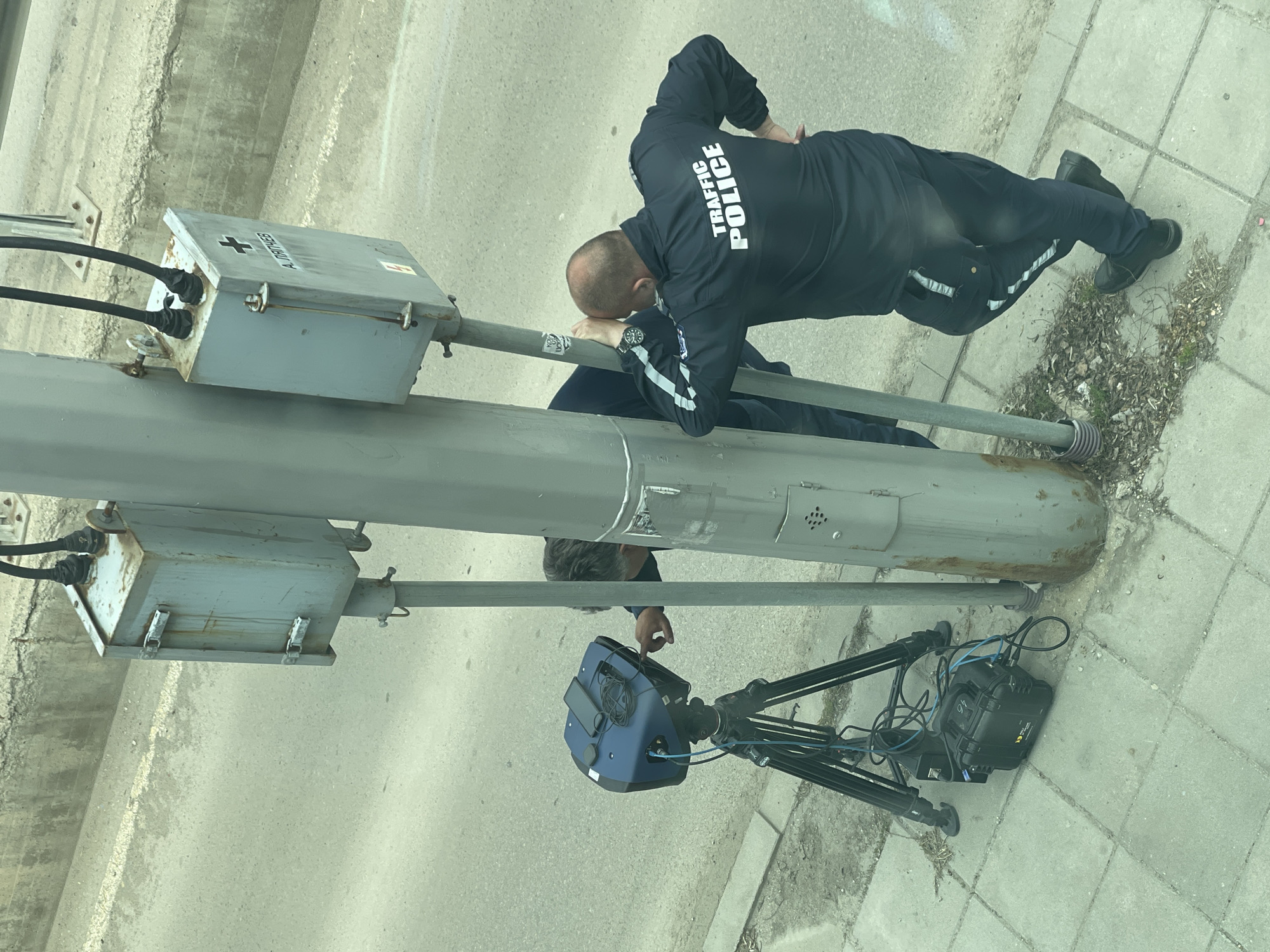The image, taken in Sofia, Bulgaria, depicts an outdoor urban setting featuring two individuals and specialized equipment. The scene is dominated by a thick, grey pole, possibly a street light or traffic signal support, alongside a paved area.
A male traffic police officer is a prominent subject, seen from behind and above. He wears a black uniform with "TRAFFIC POLICE" emblazoned in white uppercase letters across the back of his jacket. White reflective stripes are visible on his sleeves and pants. He has a watch on his left wrist and wears black shoes. The officer is leaning against the pole, his head resting on his folded arms, suggesting a moment of observation or rest.
Partially visible beneath the pole is a second person, seen from the head down to the waist, also in dark attire. This individual's hand is near a blue and black piece of technical equipment mounted on a black tripod, positioned on the concrete pavement. This equipment, likely a laser speed detection device, has various cables connected to it. Next to the tripod is a black, rugged case, possibly a power supply or data recording unit, with the number "94" visible on its side.
Two grey metal boxes are attached to the pole. The upper box has cables entering it and features the text "A. DAHNEE" and a black cross symbol on its surface. The ground consists of grey concrete paving stones with some dry vegetation. The lighting suggests it is daytime. The overall image captures a traffic police operation, likely involving speed enforcement or traffic monitoring using the specialized equipment.