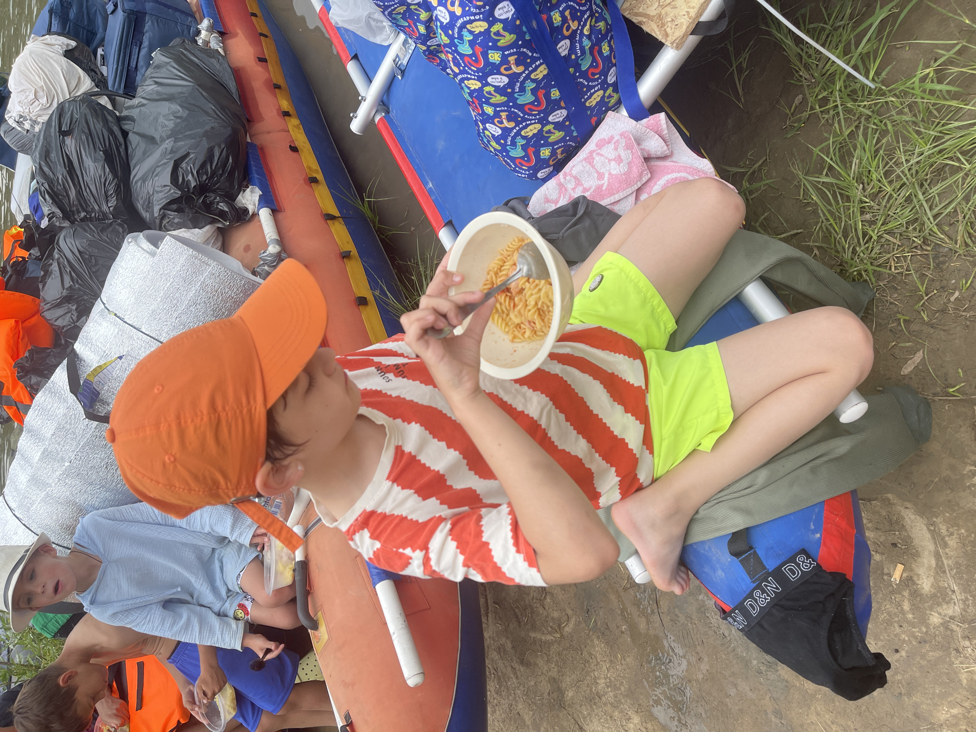 The image shows a group of children on an inflatable raft on a muddy riverbank. The central subject is a young boy wearing an orange cap and a red and white striped t-shirt, eating pasta from a bowl. To his left, another boy, wearing a white hat and a blue hooded shirt, is also holding a bowl. Several large bags, some black and some silver, are piled on the raft behind the children. The riverbank is covered in mud and sparse green grass. The overall setting appears to be outdoors, likely during a warm season. There is no indication of the location being in the city of Нежинка, Russia.
