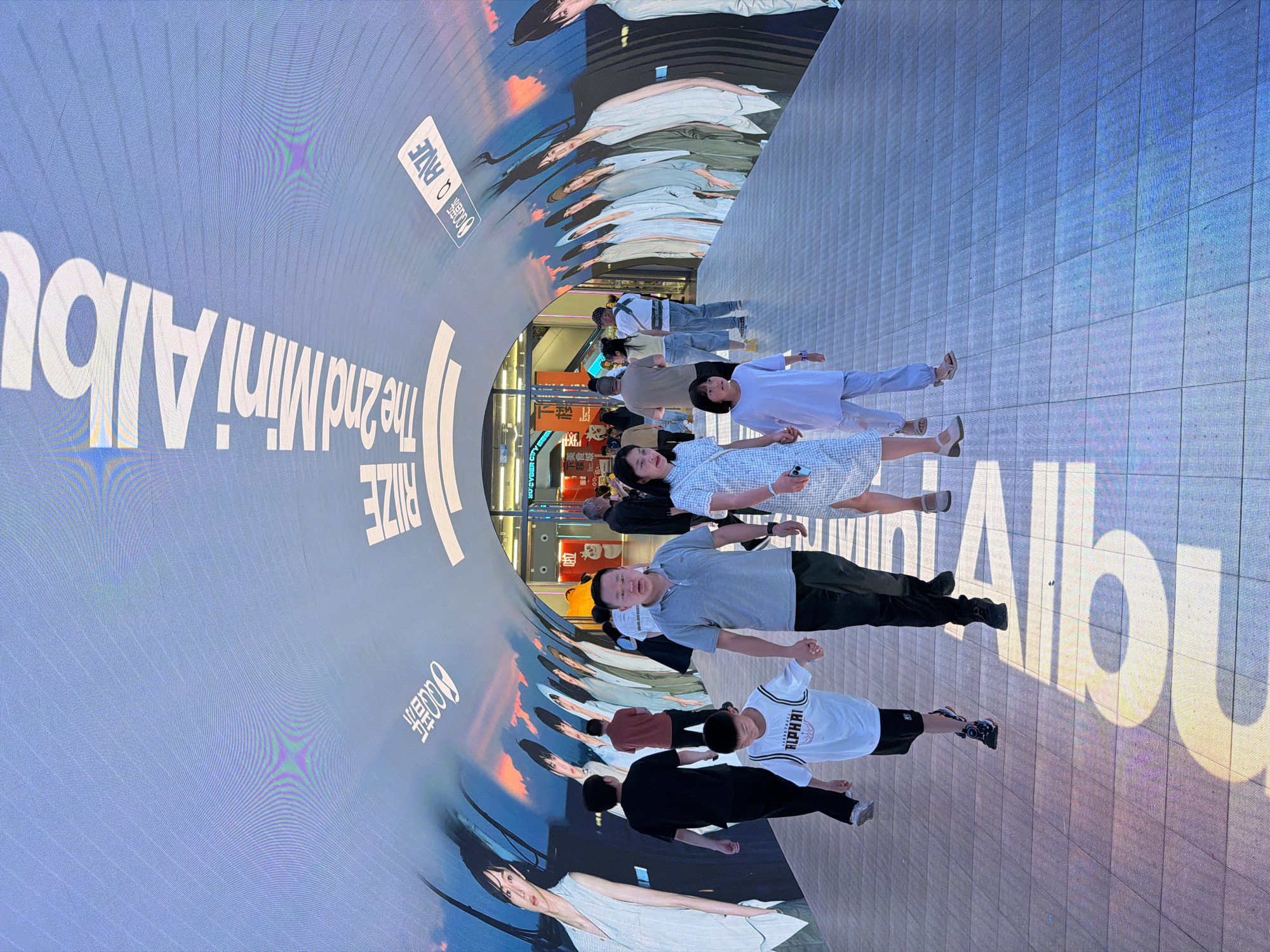 In Yuzhong District, China, a tunnel-like walkway is illuminated by a curved digital display. The display shows a series of images, including a large-scale advertisement featuring fashion models and text, possibly promoting a music album. The text "RIZE The 2nd Mini Albu" is prominently visible on the display, along with other characters that appear to be in Chinese. The walkway itself is paved with light-colored tiles and is occupied by a group of people walking in both directions. In the foreground, a family consisting of two adults and two children are walking together. The father, a man wearing a grey polo shirt and black pants, holds hands with his son, a boy in a white t-shirt with "ALPHARI" written on it and black shorts. A woman, presumably the mother, in a blue and white checkered dress, walks beside them, holding hands with a young girl in a light purple outfit. Other individuals are seen in the background, walking in the same direction as the family and in the opposite direction. The lighting suggests it is daytime, with sunlight casting shadows on the floor from the people. The overall impression is a public, pedestrian area within a modern urban setting, enhanced by digital advertising.