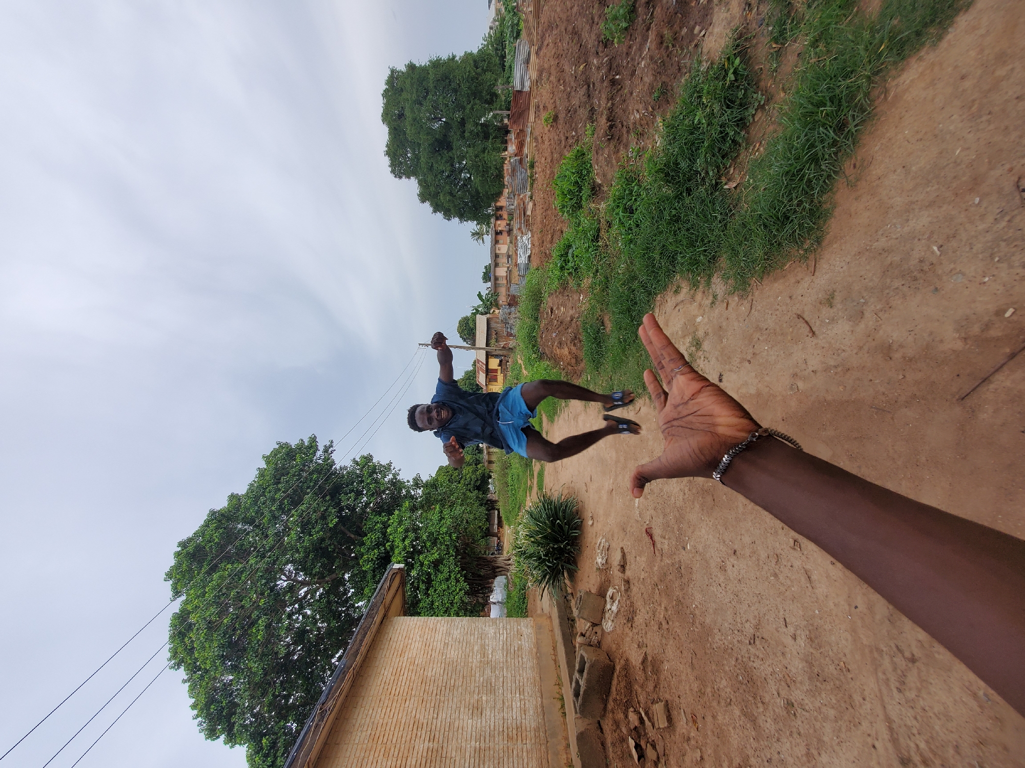 In Narayi, Nigeria, a dark-skinned man is captured mid-air, mid-jump, in front of a brick building and a large, leafy tree. He wears a blue t-shirt and blue shorts, with black flip-flops on his feet. His arms are outstretched, and he has a wide, joyful grin on his face. In the foreground, a dark-skinned arm, adorned with a silver bracelet, is extended upwards, as if reaching for the jumper. The ground is composed of reddish-brown dirt, with patches of green grass and scattered debris, including concrete blocks. The background reveals more of the urban landscape, with other buildings, corrugated metal structures, and more trees under a cloudy sky. The overall scene conveys a sense of playful interaction and exuberance in a rural or semi-urban Nigerian setting.