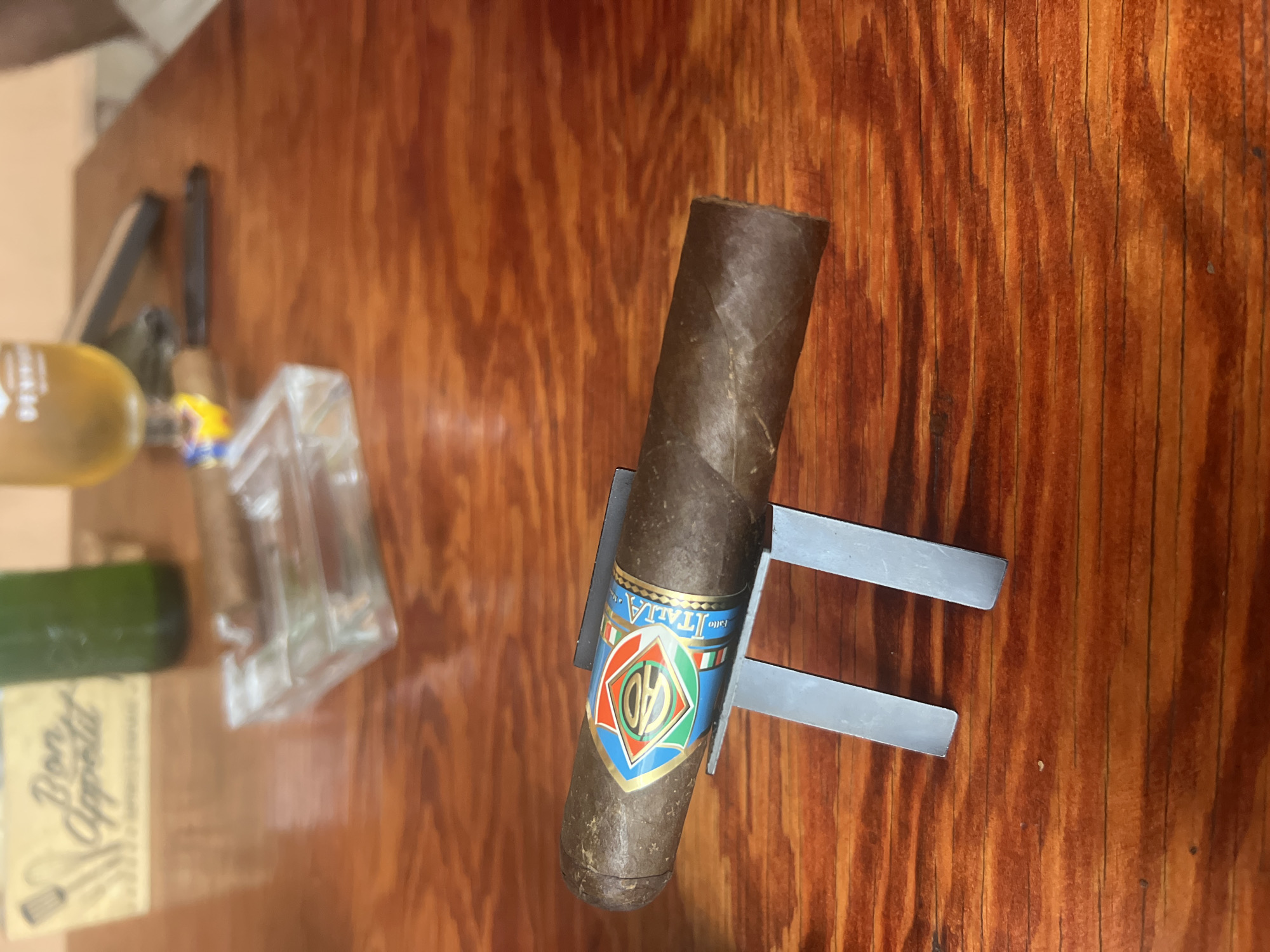 The image depicts an indoor scene in Sofia, Bulgaria, featuring a dark brown cigar prominently displayed on a metallic stand. The cigar has a textured wrapper and is adorned with a colorful band featuring a central diamond motif in red, green, blue, and gold, enclosing the letters "CAO," with "ITALIA" written vertically along its right side. The stand is composed of two vertical, parallel silver-colored metallic bars supporting the cigar horizontally.
In the blurred background, a polished reddish-brown wooden table surface with distinct grain patterns serves as the base for various items. To the left, a light-colored rectangular wooden sign is visible, bearing the cursive text "Bon Appetit" above silhouettes of kitchen utensils. Behind this sign, a dark green glass bottle stands upright. To the right, a clear glass ashtray holds a lighter-colored cigar in one of its indentations. Further right, a blurred yellowish glass, possibly a beverage glass, is present, with indistinct dark objects nearby on the table. A faint, blurred light-colored shape on the far right edge of the frame suggests the presence of a person. The scene is well-lit, indicating an ambient indoor environment.