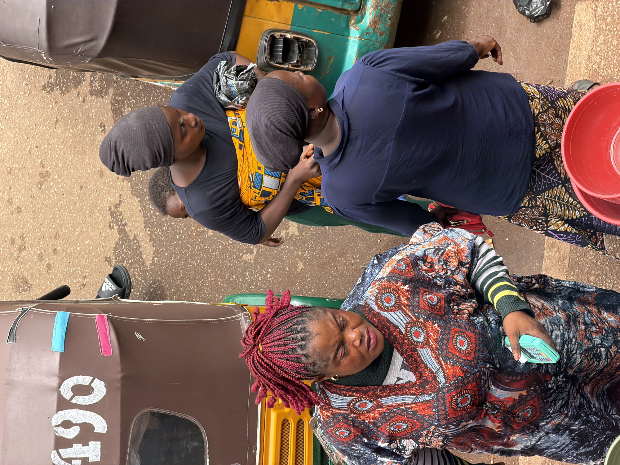 In Bukuru, Nigeria, a scene unfolds at a roadside with two auto-rickshaws, one brown and yellow, the other green and yellow, parked close together on a dusty, paved surface. Three women and a child are the central figures.
The woman in the foreground, with vibrant red braided hair, wears a patterned, colorful dress over a striped long-sleeved shirt. She holds a small, turquoise electronic device in her right hand. To her left, and slightly behind, a woman in a dark blue long-sleeved top and a patterned skirt stands looking away from the camera, her left arm extended slightly.
Behind them, a woman with a black headscarf, dressed in a black t-shirt and an orange and yellow patterned wrap, is visible from the chest up. A small child's head with curly hair peeks out from behind her. The child appears to be looking forward.
The brown auto-rickshaw has the number "490" stenciled in white on its side, with three colorful fabric tabs – blue, pink, and black – attached near the top. A person's feet wearing black Adidas slides are visible near the back of this rickshaw. The green and yellow auto-rickshaw has a broken headlight housing on its side. The ground is a mix of reddish-brown earth and scattered debris. Two red plastic buckets are prominently placed in the lower right corner of the image.