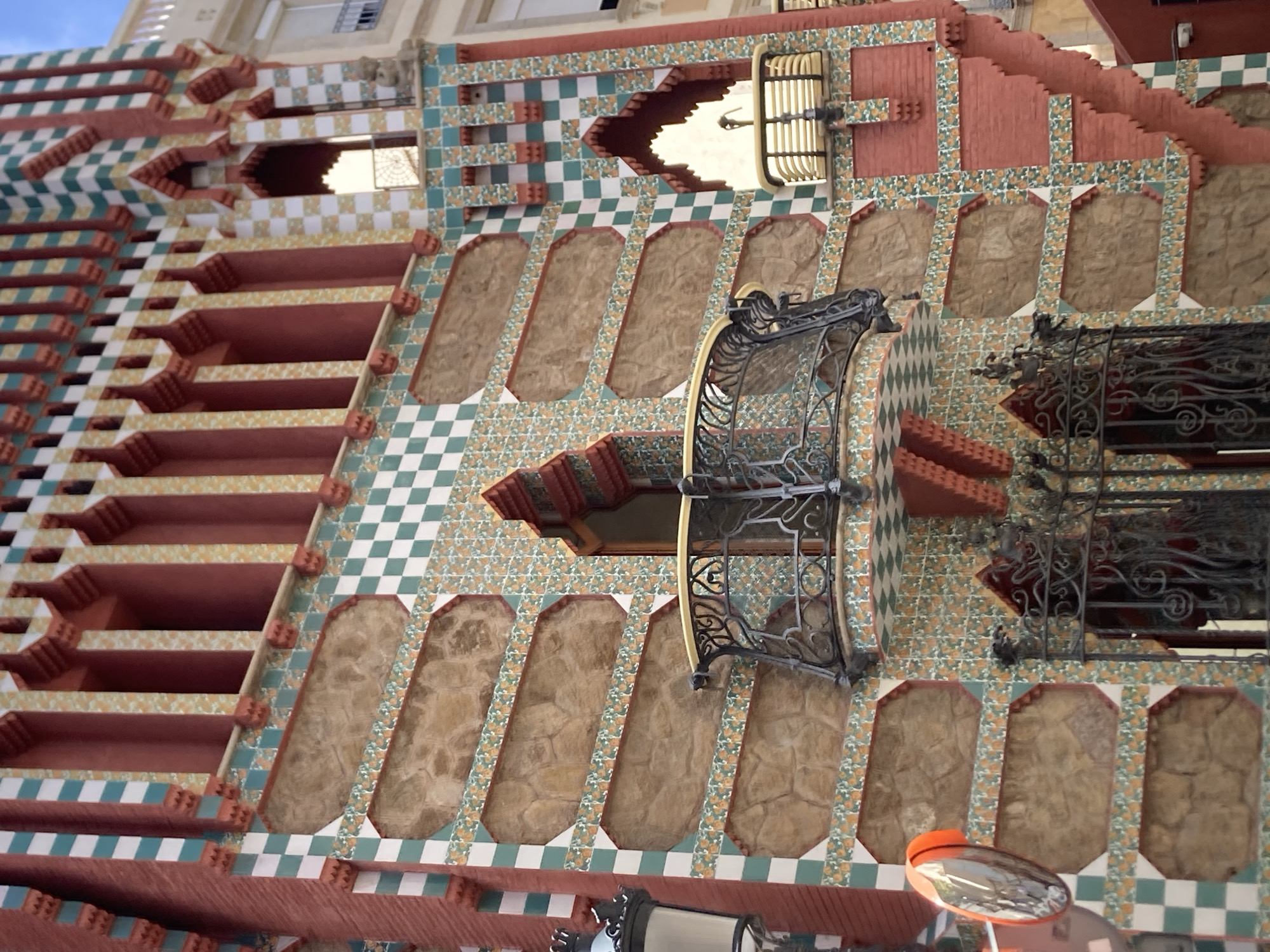 The image shows a close-up of the exterior of Casa Vicens, located in the Gràcia district of Barcelona, Spain. The building features a distinctive facade with a combination of red brick, green and white checkerboard tiles, and stone-like textured panels. Ornamental details include red columns with decorative tops and intricate wrought-iron balconies and grilles. A convex mirror with an orange frame is visible on the left side of the image. The architectural style appears to be early Modernisme, characterized by vibrant colors and decorative elements inspired by nature.