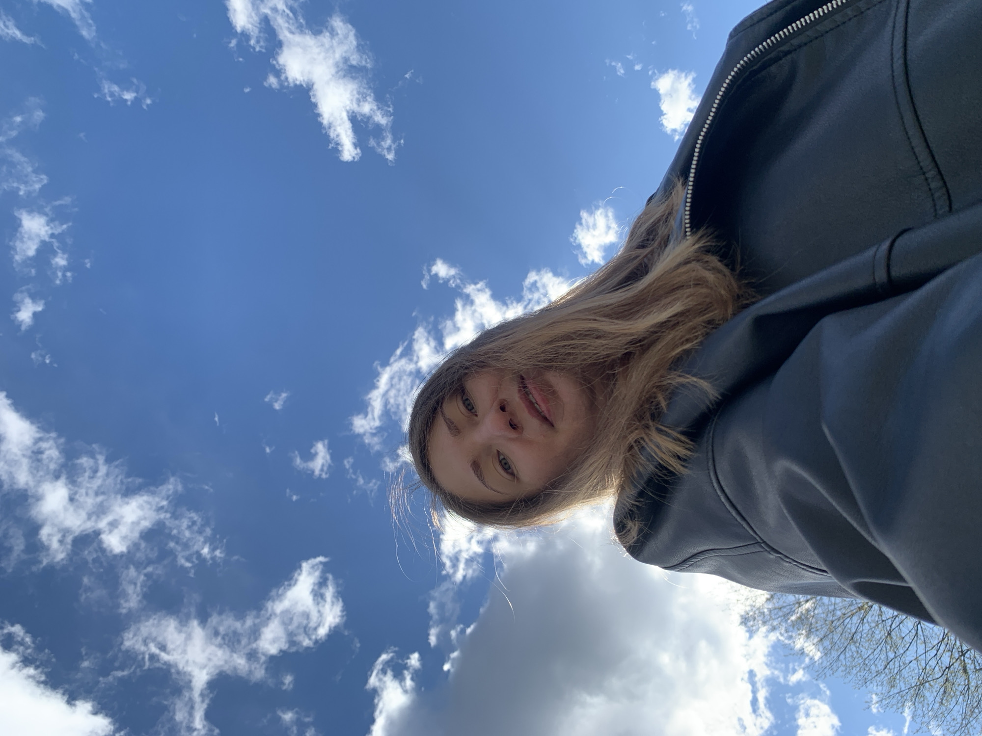 A low-angle selfie was taken outdoors during the day. A fair-skinned person with blonde hair is wearing a black leather jacket. The background is a bright blue sky with scattered white clouds. The location might be Chaikovsky, Russia.