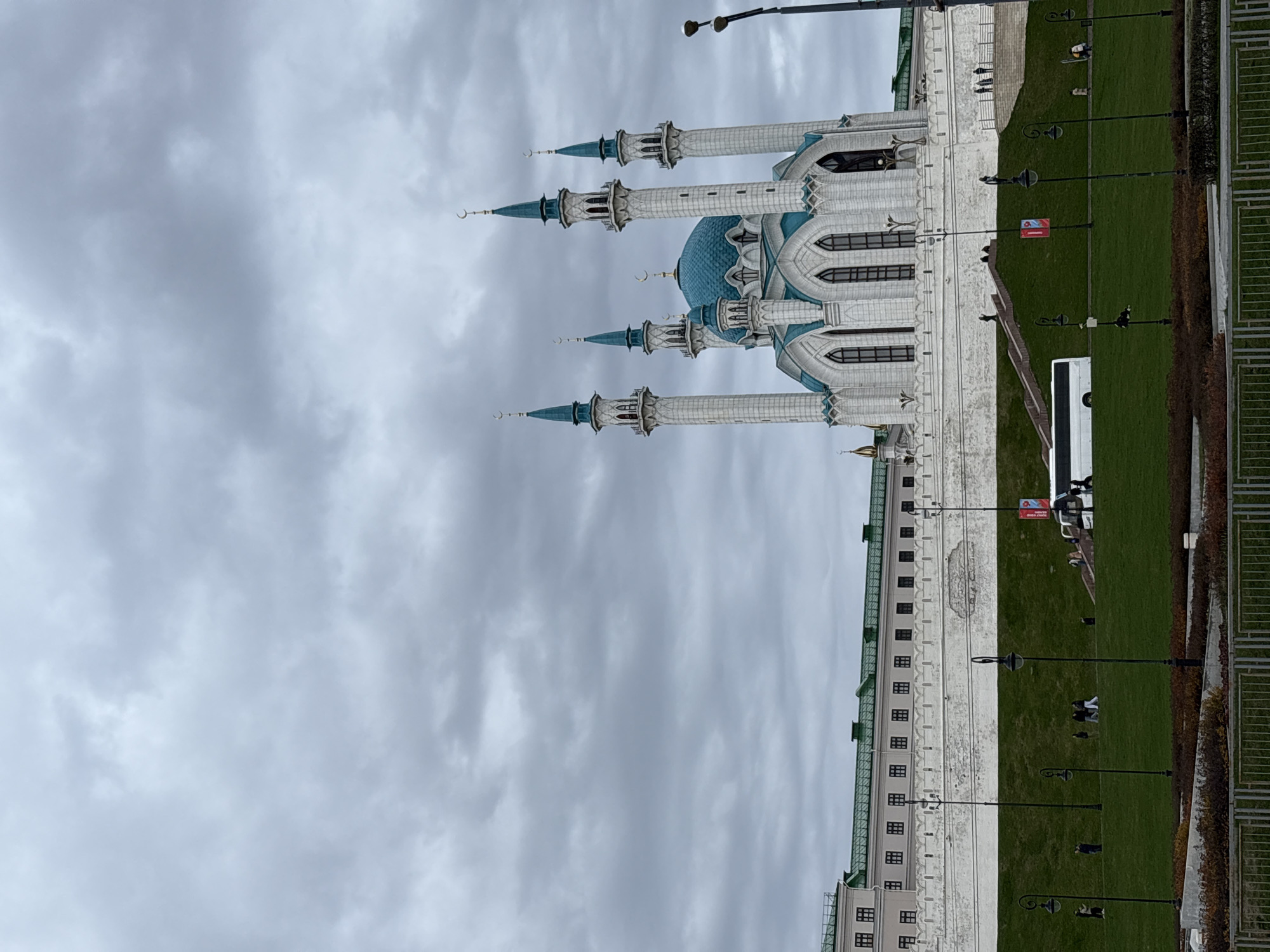This media file captures an exterior view of the Kul Sharif Mosque in Kazan, Russia, under an overcast sky. The image is taken from a slightly elevated perspective, looking towards the mosque and its surrounding grounds.
The Kul Sharif Mosque is the central subject, a magnificent structure with white and light blue architectural details. It features a large, fluted blue dome crowned with a golden crescent moon, flanked by four prominent minarets, each topped with a smaller blue dome and a golden crescent. Two additional, smaller minarets are visible on either side of the main entrance. The mosque's walls are intricately decorated with arched windows and patterned stone work.
To the right of the mosque, a wide expanse of neatly manicured green grass stretches across the frame. Several black ornamental lampposts are spaced throughout this grassy area. In the foreground on the right, a white bus is parked, and a few people are visible on the grass, some walking, others standing. More individuals are seen closer to the mosque, near a set of steps leading up to the entrance.
In the background, behind the grassy area and to the left of the mosque, a long, light-colored building with a green roof and numerous small, dark windows is visible, likely part of the Kazan Kremlin complex. The sky is uniformly grey and cloudy, suggesting an overcast day, without clear sunshine. On one of the lampposts, a red banner is affixed, bearing the Cyrillic word "Татнефть" (Tatneft), a Russian oil company.