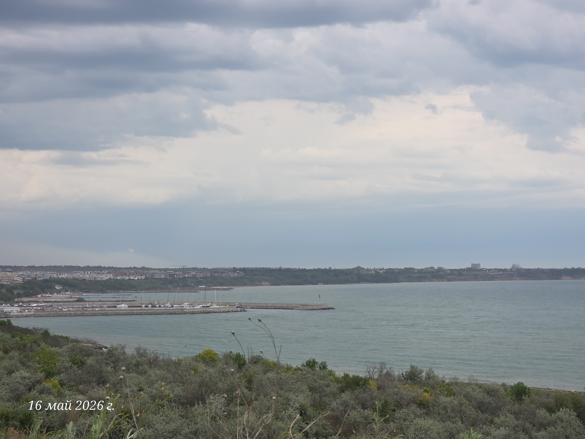 The image depicts a coastal scene in Burgas, Bulgaria on May 16, 2026. The foreground consists of low-lying, scrubby vegetation. Beyond the vegetation lies the Black Sea, calm and grey under an overcast sky. A marina with docked boats is visible along the coastline, protected by a breakwater. In the distance, a skyline of buildings is present along the coast. The overall atmosphere is muted and subdued due to the weather conditions.