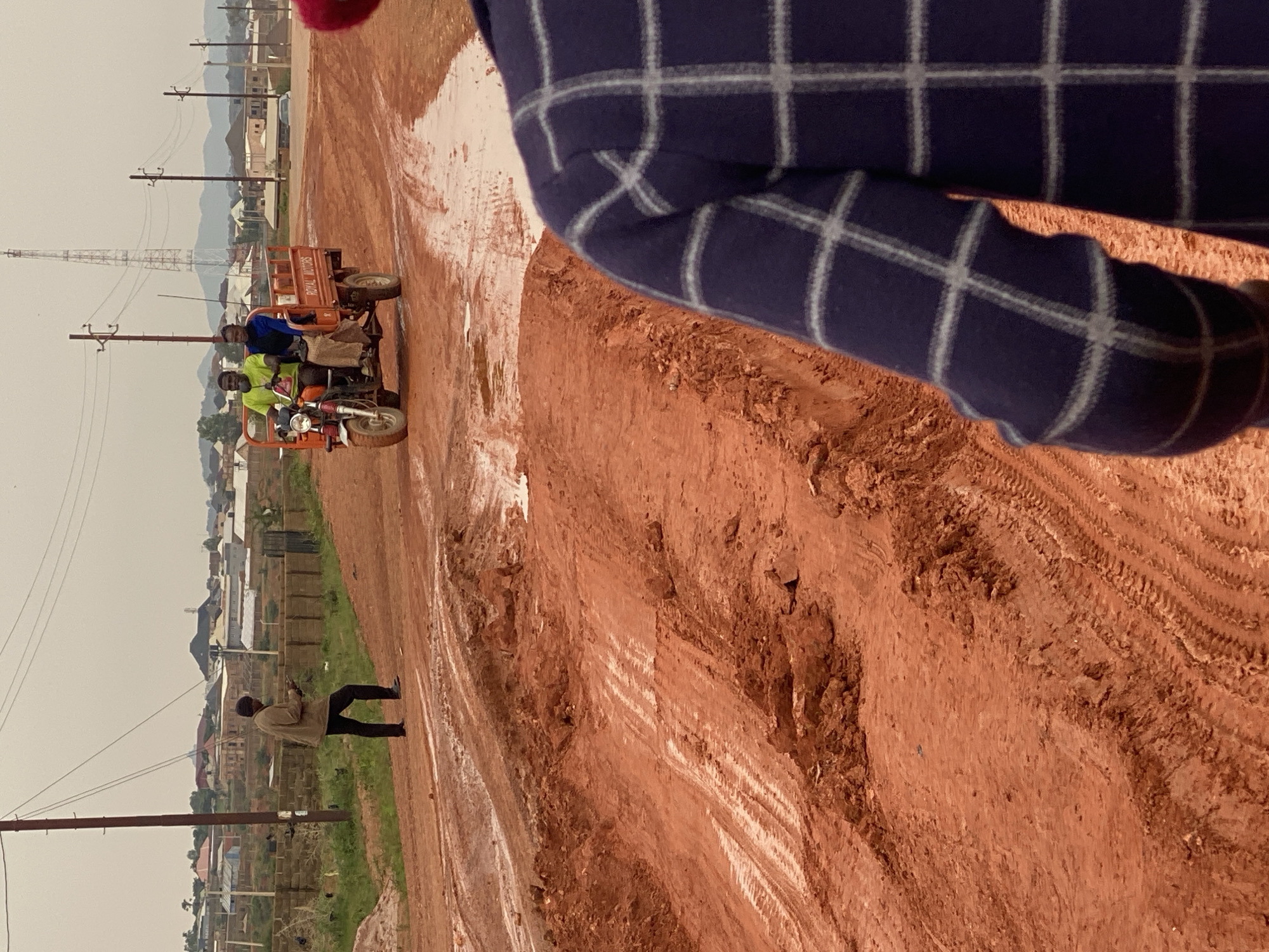 An overcast day in Jos, Nigeria, shows a dirt road under construction. In the foreground, the back of a person wearing a blue and white checkered shirt is visible. Further down the road, a person in a brown jacket and dark pants stands on the left, looking at their phone. On the right, an orange tricycle with three occupants drives along the muddy road. The tricycle is labeled "ROYAL MOTORS". In the background, houses and a radio tower are visible, suggesting a semi-urban or developing area. The ground is red, indicating the soil type of the region.