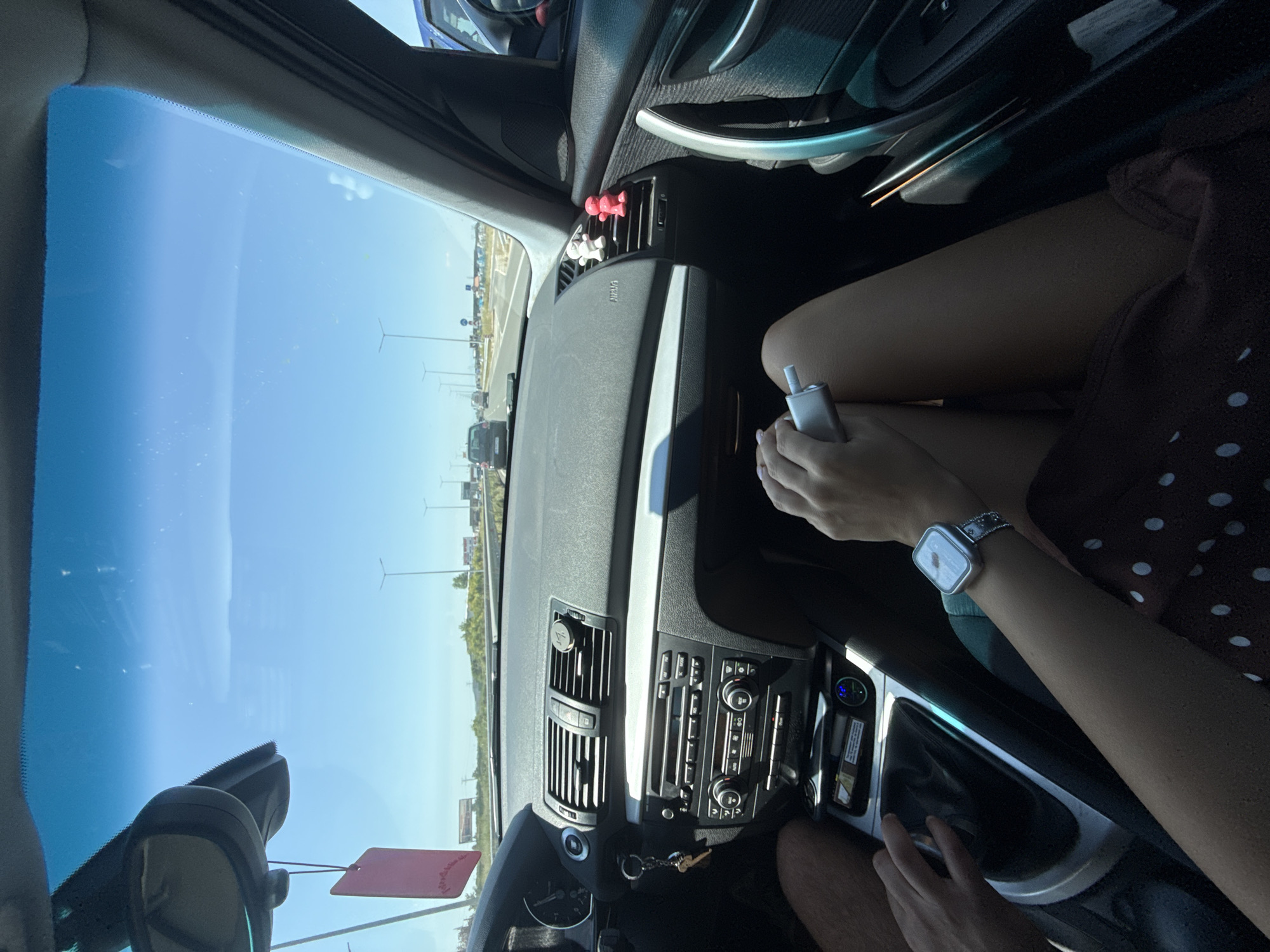 The image depicts the interior of a car, looking forward through the windshield on a sunny day. The perspective is from the passenger seat. In the foreground, a person's legs are visible, wearing a dark red dress with white polka dots. The person is holding a silver electronic cigarette device in their right hand and wearing a silver smartwatch with a white face and silver band on their left wrist. To the left, a driver's hand is visible resting on the gear shift.
The car's dashboard is prominent, featuring a car stereo system with various buttons and knobs, air vents, and a steering wheel on the left. A pink air freshener hangs from the rearview mirror. Two small, brightly colored figures, one red and one white, are attached to the dashboard near the passenger-side air vent.
Outside the car, the view shows a highway with multiple lanes of traffic. Several streetlights are visible along the roadside, as are some trees and distant billboards. The sky is clear and blue. The location is identified as Krumovo, Bulgaria.