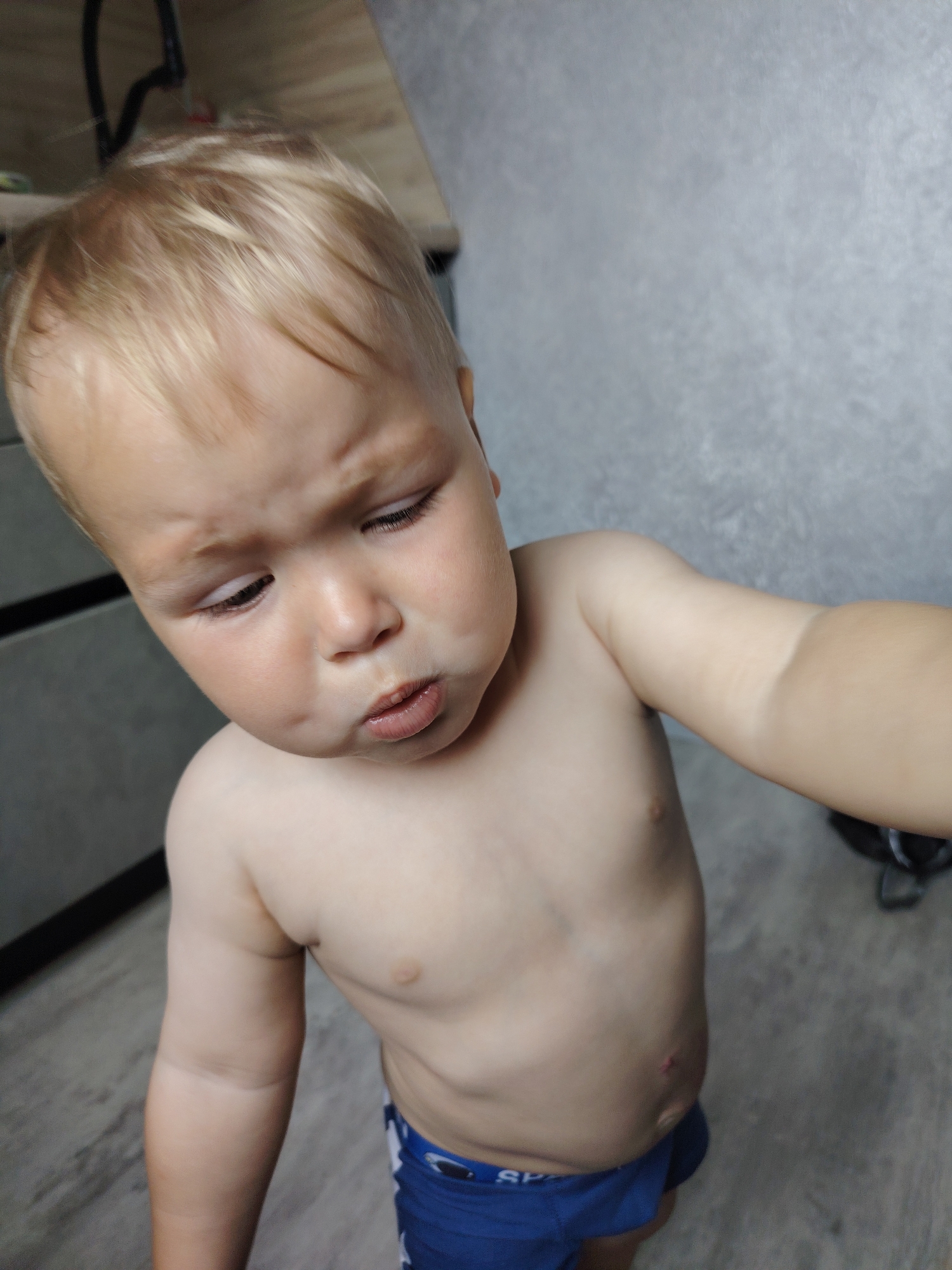 A toddler with blond hair is looking down with a curious expression. The child is shirtless, wearing blue shorts. The background shows a grey textured wall and wooden furniture. The image was taken indoors. No specific location in Chaikovsky, Russia can be determined from the image.