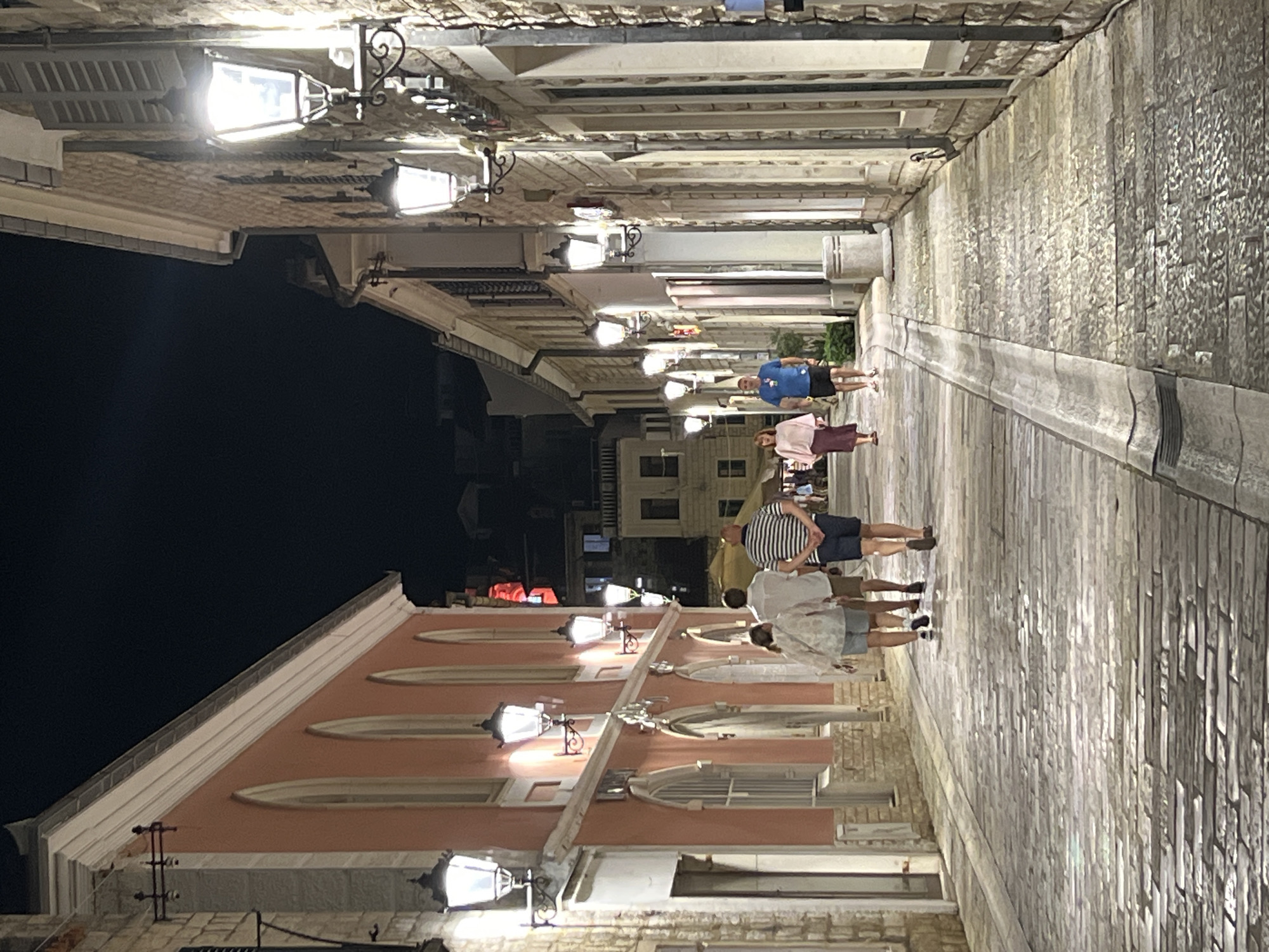 In Herceg Novi, Montenegro, a narrow, cobblestone street is illuminated by old-fashioned street lamps at night. The street is lined on both sides with buildings. On the left, a salmon-pink building features arched windows, and on the right, a stone building has traditional shutters. Several people are walking along the street. In the foreground, a group of three individuals walks away from the camera. Further down the street, two people are walking towards the camera. The lighting from the lamps casts long shadows on the wet-looking cobblestones.