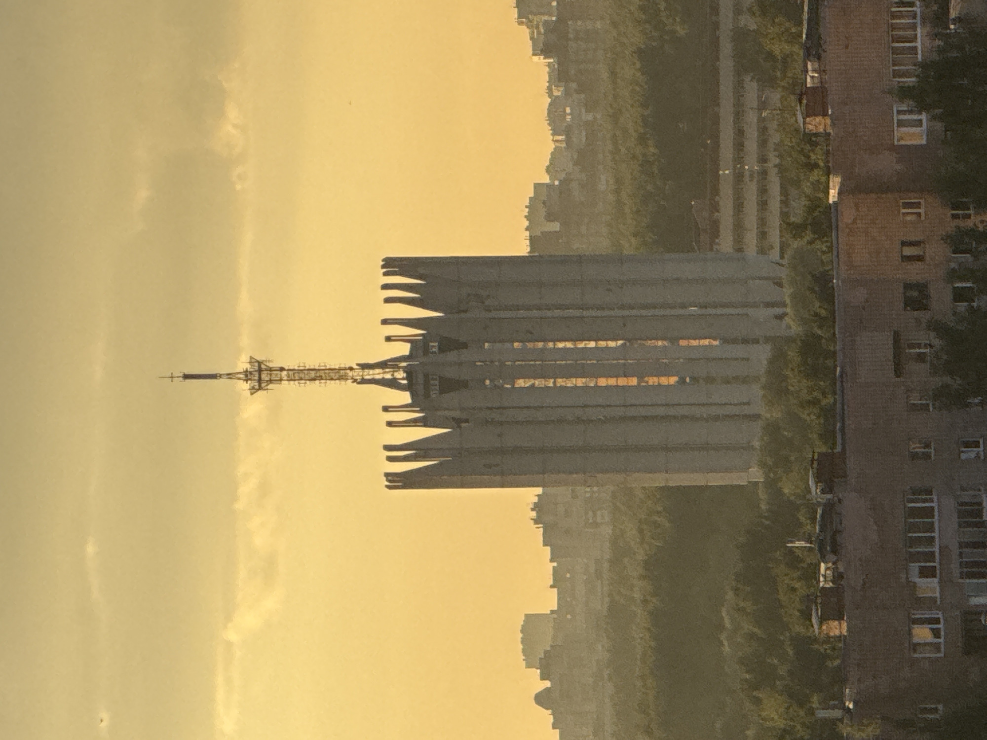 The image captures a city skyline at sunset, bathed in a warm, golden light. A prominent, uniquely shaped concrete tower with a spire on top dominates the center of the frame. The tower is surrounded by a dense forest, with a sprawling cityscape of buildings visible in the background. In the foreground, a brick apartment building is partially visible. The sky is a soft yellow with scattered clouds. The overall scene suggests a tranquil urban environment as day transitions into evening.