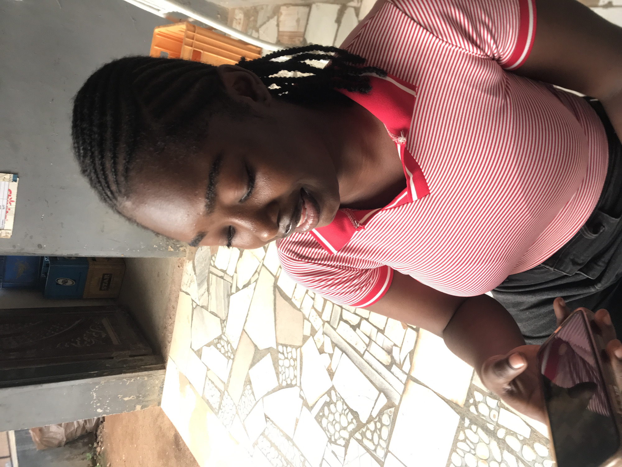 A medium shot, taken during the daytime, captures a person with dark skin and dark braided hair, looking down at a smartphone with a gentle smile. The person wears a red polo shirt featuring thin white horizontal stripes, a red collar with a white stripe, and white trim on the sleeves, paired with dark trousers or jeans. Their eyes are cast downwards, indicating focus on the phone held in their hands.
The setting appears to be an indoor or semi-enclosed outdoor area, possibly a patio or entryway. The floor is covered in a decorative mosaic-like pattern of irregularly shaped light-colored tiles, with some sections incorporating smaller, dark, pebble-like elements. In the background, a dark wooden doorway is visible, behind which are stacks of plastic crates. Text on a blue crate partially reads "NBA", and on a yellowish crate, "STAR" is discernible, suggesting beverage brands common in Nigeria. The surrounding walls are a weathered light grey. The person's expression conveys a sense of quiet engagement or contentment. The location context is New Stadium, Nigeria.