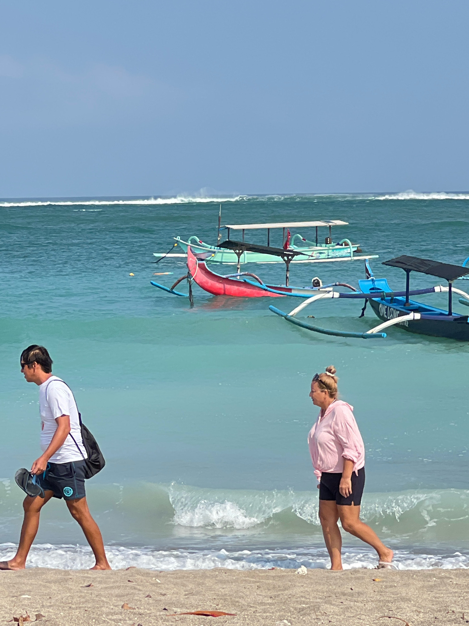 Two people walk on the beach in Kuta, Indonesia, with traditional Balinese fishing boats anchored in the turquoise water behind them. The man on the left is wearing a white t-shirt and dark shorts, carrying a bag over his shoulder and flip-flops in his hand. The woman on the right is wearing a pink hooded shirt and black shorts. In the background, several colorful boats with outriggers are visible, bobbing on the gentle waves. The sky is a clear blue, and the water is a vibrant aquamarine color, indicative of the tropical climate. The beach itself is sandy with some debris scattered on the shore.