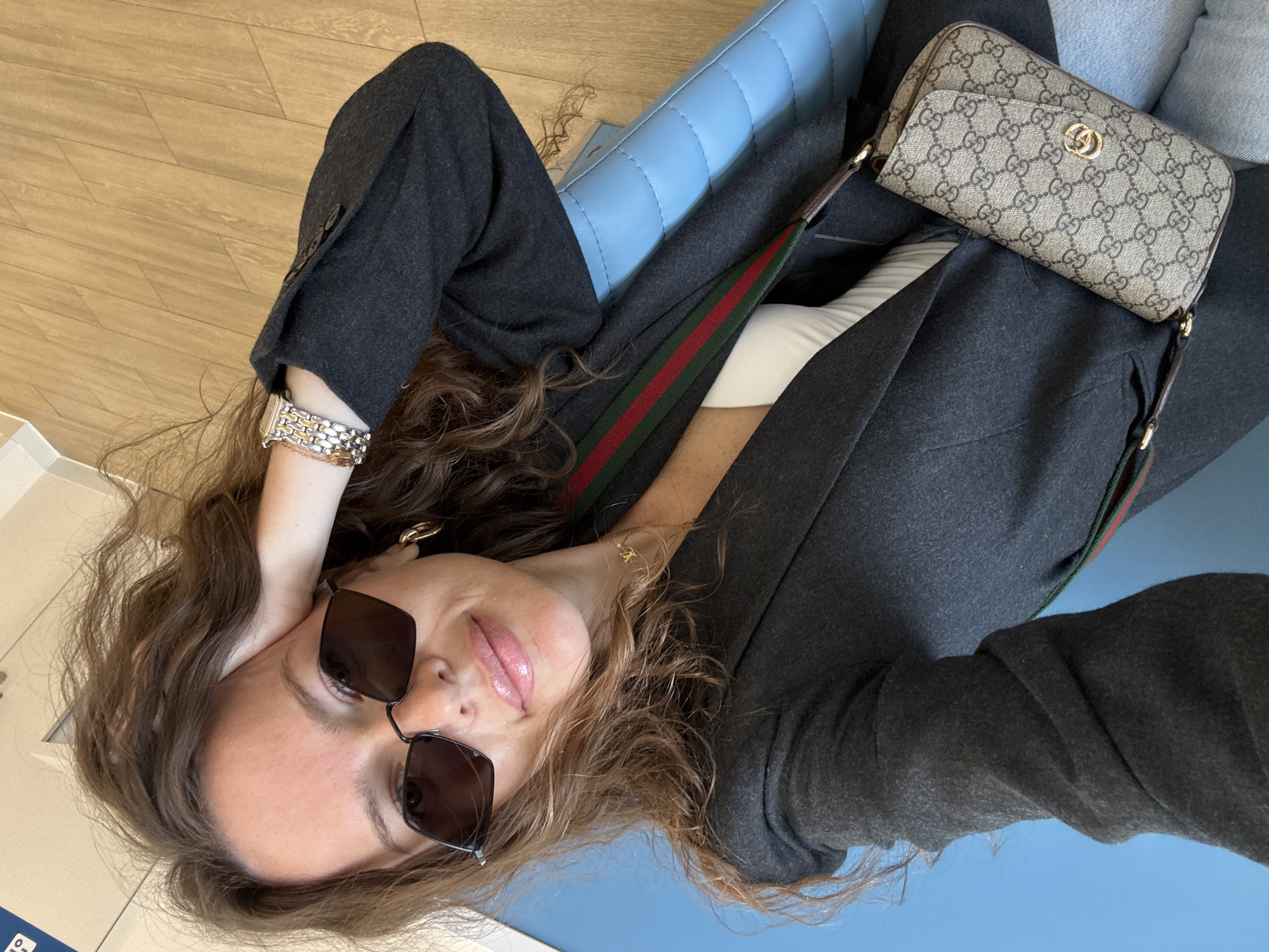 In Moscow, Russia, a woman with curly brown hair and sunglasses is captured in a close-up shot. She is wearing a dark gray blazer over a white top, and a crossbody bag with the Gucci logo is slung across her. A distinctive red and green stripe adorns the strap of the bag. Her right hand rests on her cheek, and she is adorned with earrings and a watch on her left wrist. The background features a bright blue surface, possibly a chair or wall, and a wooden floor. The lighting suggests an indoor setting with natural light contributing to the scene.