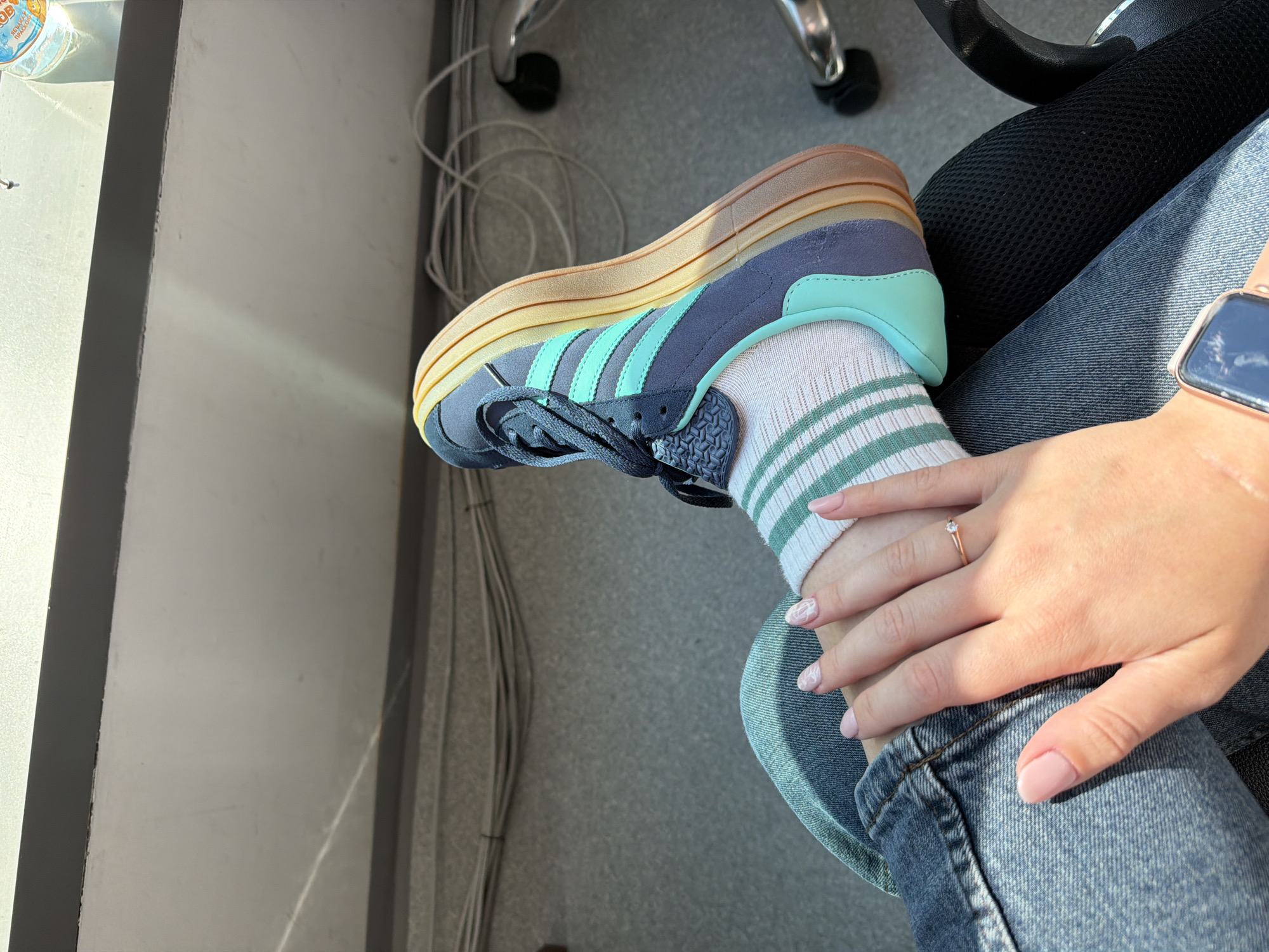 The image shows a person's left leg and foot, wearing a blue and turquoise Adidas sneaker with a multi-layered, gradient-colored sole. The person is also wearing light blue denim jeans and white socks with green stripes. A hand with light pink nail polish is resting on the jeans. The background appears to be an indoor environment, possibly an office, with gray carpeting, electrical cords, and the edge of a chair. The scene is lit by sunlight casting shadows. There is no indication of the city of Sofia, Bulgaria, in the image.