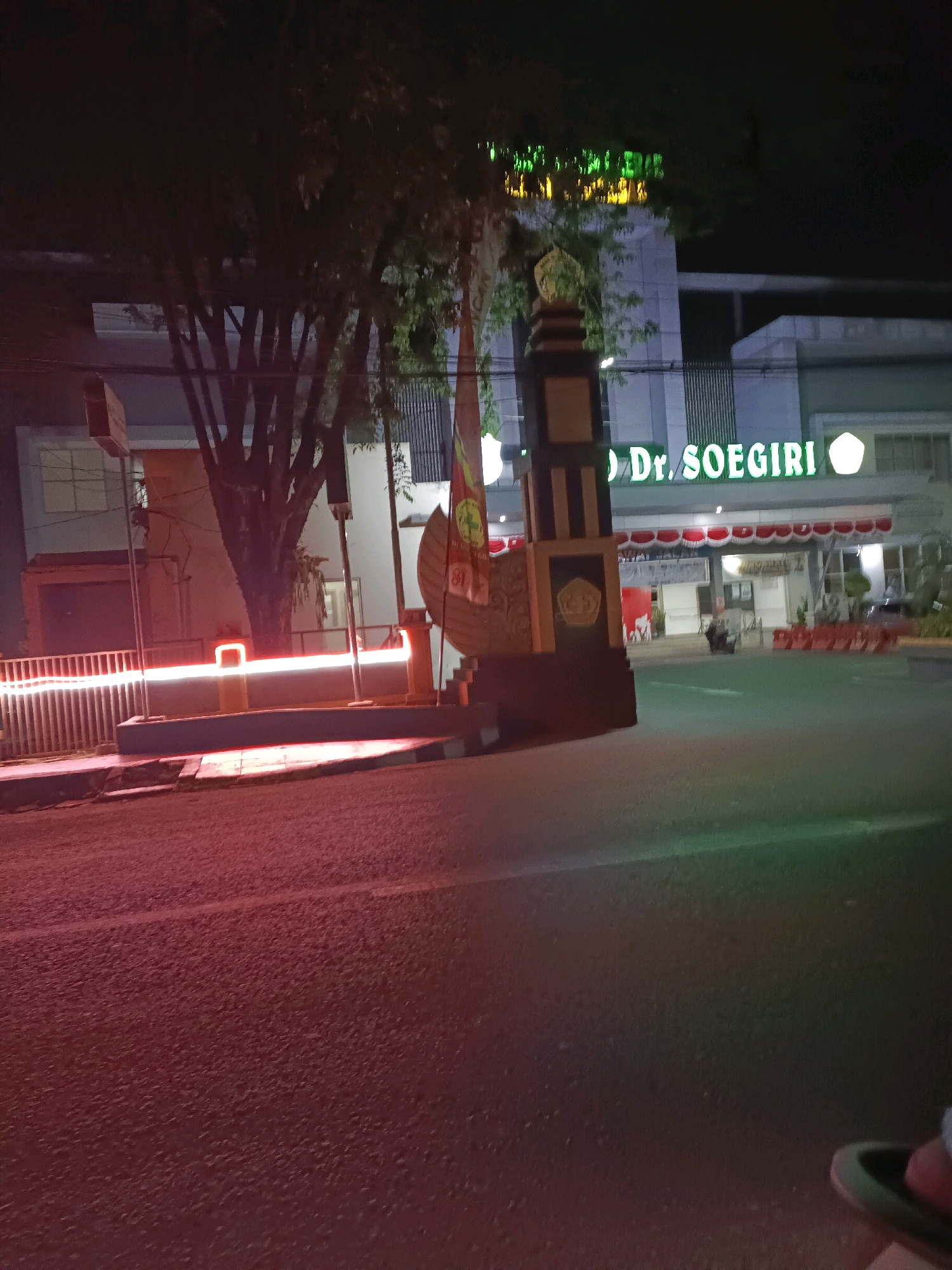 The image shows the Dr. Soegiri Lamongan Hospital entrance at night. A large illuminated sign above the entrance reads "Dr. SOEGIRI" in green neon letters. In front of the hospital, there is a decorative monument with flags and emblems. Red and white awnings are visible above the entrance. The street in front of the hospital is paved and illuminated by red neon lights from a railing system. Trees are present on the left side of the image, casting shadows. The scene is lit by artificial lighting, indicating it is nighttime.