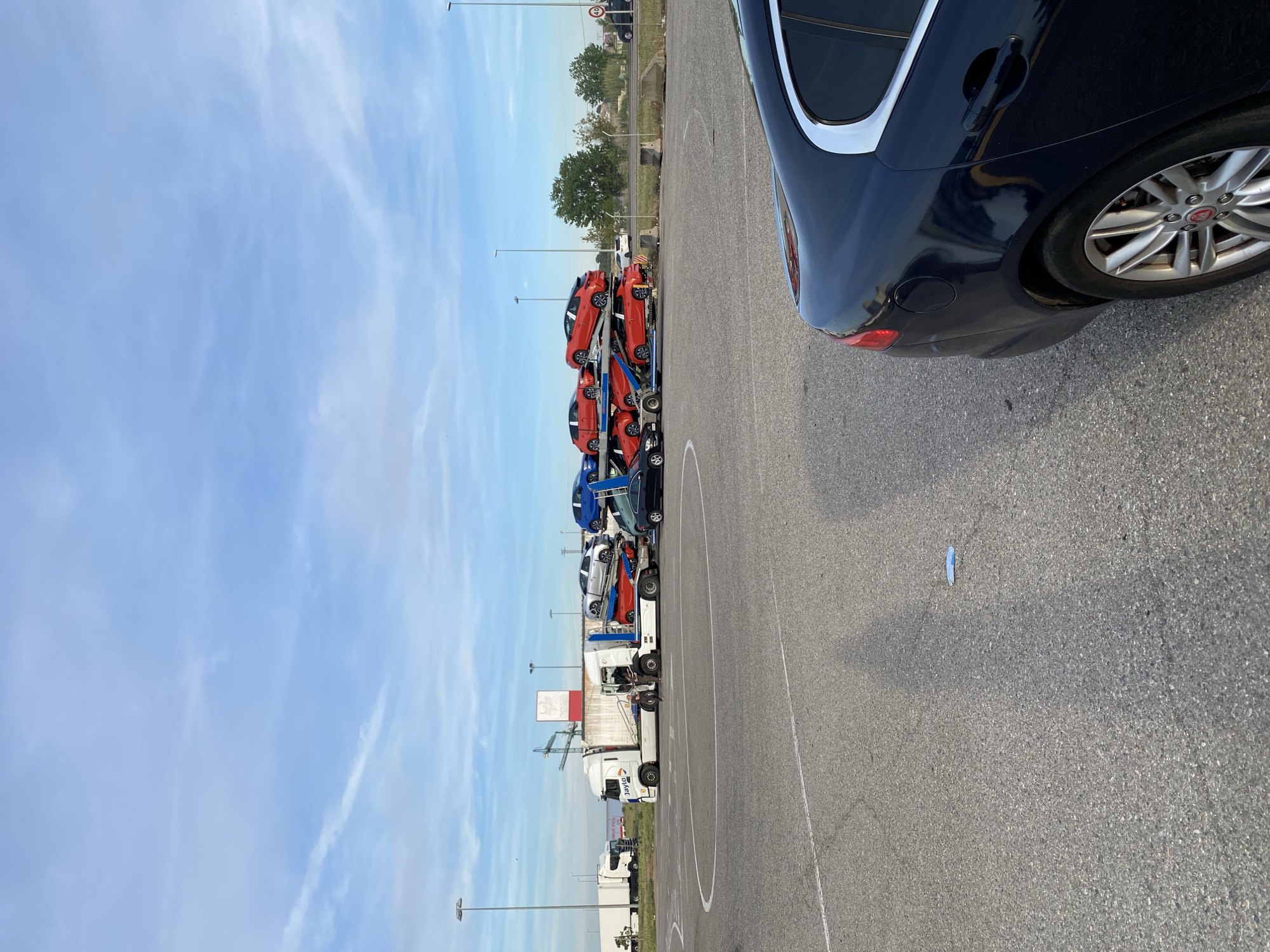 The image depicts a parking lot in Lleida, Spain. A dark-colored car occupies the right side of the frame, partially obscuring the background. In the distance, a car carrier truck is parked, loaded with multiple new cars of various colors, primarily red and blue. The sky is light blue with scattered clouds. Streetlights are visible in the background, along with some greenery and buildings. The parking lot is paved with asphalt and marked with painted lines. A small piece of blue debris is visible on the asphalt surface.