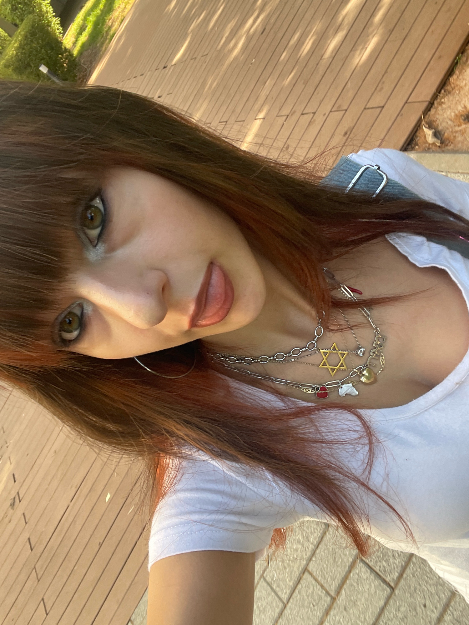 A young person with auburn hair and bangs is taking a selfie outdoors. They are wearing a white t-shirt and multiple necklaces, including one with a prominent yellow Star of David pendant. Their left eye is visible, with green irises and heavy eye makeup. Their right hand is partially visible, as are their lips, which are slightly parted. The background consists of a wooden deck and some greenery, suggesting a public park or outdoor seating area. The lighting indicates it is daytime, possibly late afternoon due to the shadows. The orientation of the photo is sideways. The location is identified as Raanana, Israel.