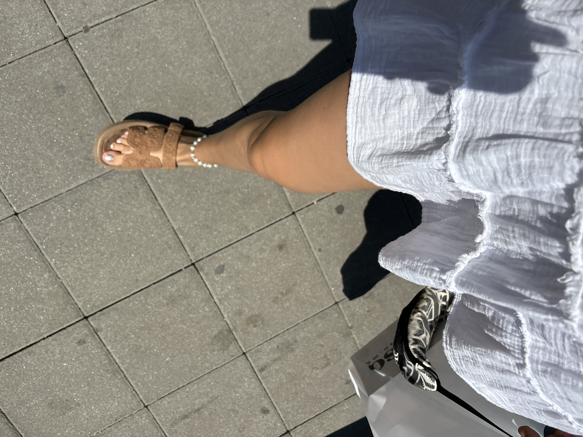 A person is walking on a sidewalk in Budva, Montenegro, captured from a high-angle perspective. The sidewalk is paved with gray square tiles. The person is wearing a white, crinkled fabric dress and brown fuzzy sandals with a butterfly design. They are also wearing a delicate pearl anklet. In the left of the frame, a white shopping bag with black lettering is visible, with a black and white patterned item peeking out of it. The sun is casting a strong shadow of the person onto the sidewalk. The overall scene suggests a sunny day in an urban environment.