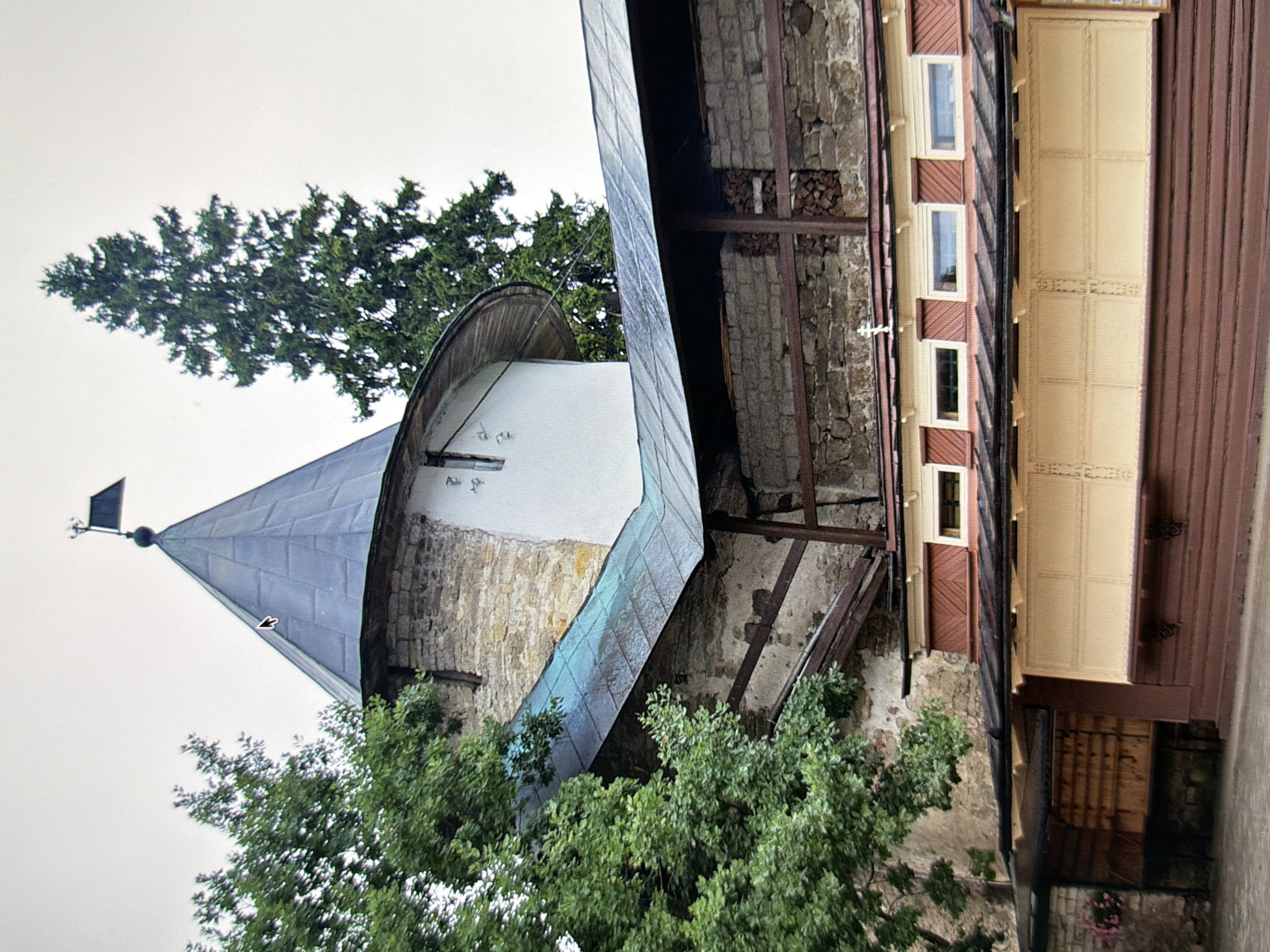 The image depicts a section of the Kyiv Pechersk Lavra, a historic Orthodox Christian monastery in Kyiv, Ukraine. In the foreground, a low-lying structure with a reddish-brown wooden facade and light-colored window panels is visible. Above this, a stone wall is partly covered by a slanted, blue-tiled roof supported by wooden beams. Further back, a white cylindrical tower with a conical, dark grey metal roof rises, topped by a dark flag on a pole. A large coniferous tree stands to the right of the tower, and lush green foliage from other trees partially obscures the left side of the tower. The sky is overcast. The architecture suggests a blend of historical and functional elements within the monastery complex.