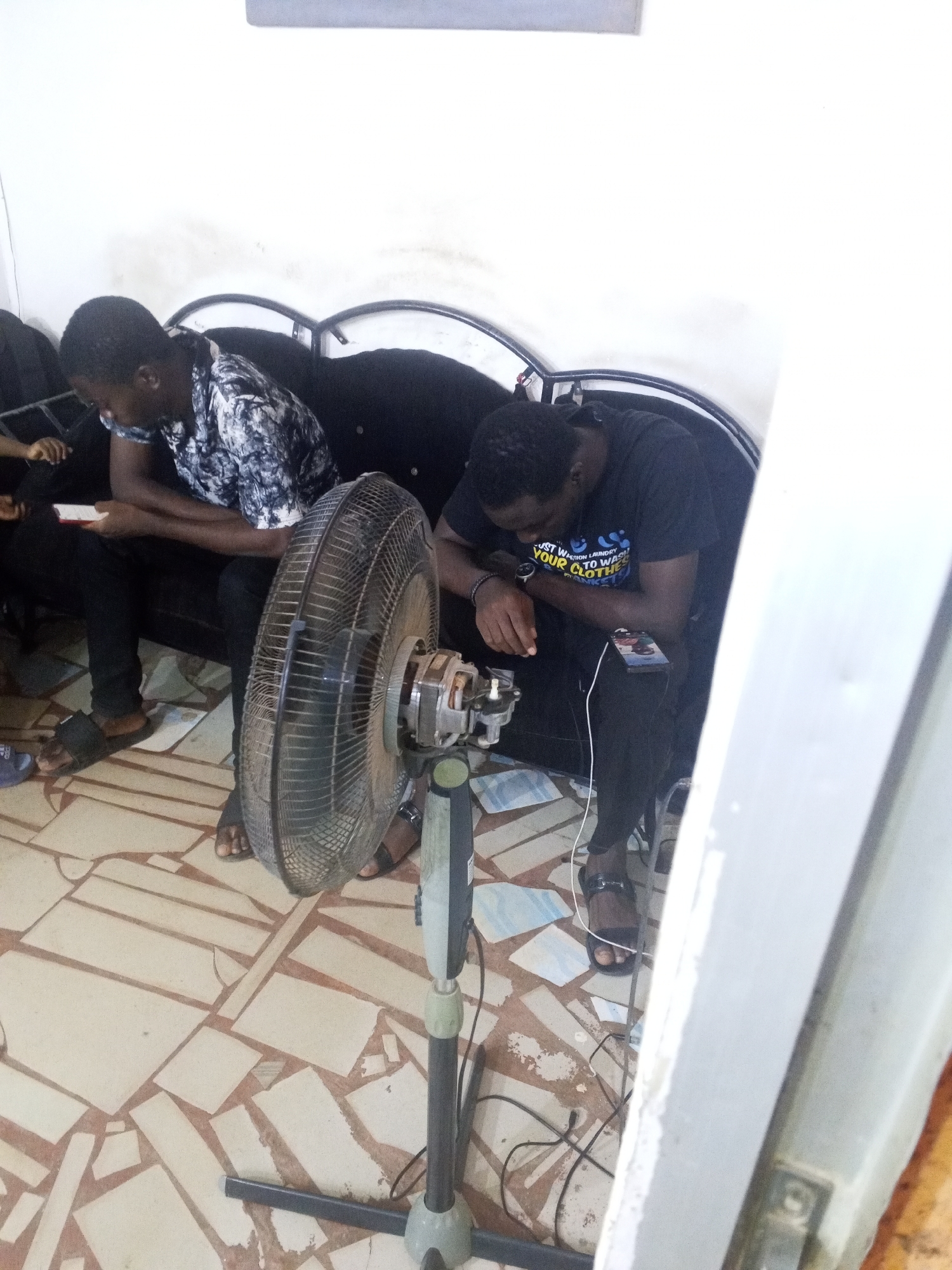 Two young men are seated on a black couch. One is wearing a black and white patterned shirt and black pants, looking at a phone. The other man wears a black t-shirt with a colorful graphic and black pants, with his head bowed. A standing fan is in front of them on a tiled floor. The location is in Gwong, Nigeria.