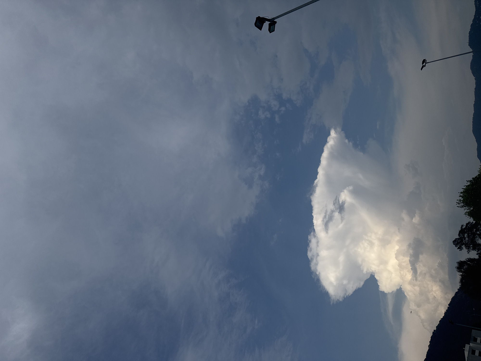 The image depicts a dramatic sky over Chiasso, Switzerland, dominated by a large, towering cumulonimbus cloud, often referred to as a thunderhead, which has developed an anvil-like shape at its top. The cloud is brightly lit from above, indicating sunlight, while its base is darker, suggesting density and potential precipitation. The sky is a mixture of deep blue in some areas and overcast gray clouds in others, creating a sense of atmospheric depth and dynamism. In the foreground, silhouetted against the sky, are the dark shapes of trees and hills, indicating a natural landscape on the outskirts of the city. Two modern streetlights with multiple lamps are visible, one on the right side and another further in the distance towards the bottom right, suggesting an urban or semi-urban environment. A small, dark object, possibly an airplane or bird, is visible in the sky to the left of the large cloud. A building facade is partially visible in the bottom left corner. The overall scene is one of natural power and the interplay of light and shadow in a meteorological event.