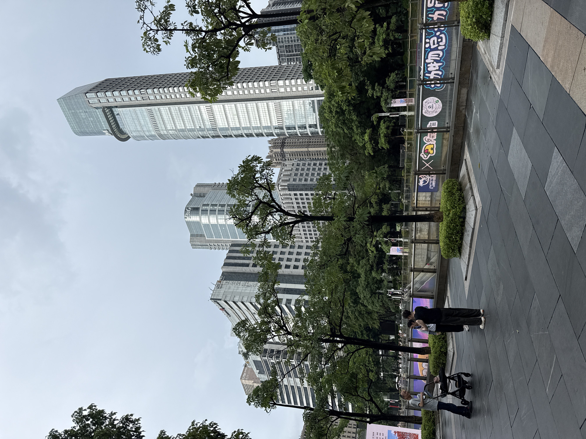 This media file captures an outdoor scene in Tianhe District, China. The primary subjects are several people interacting, with two individuals standing close together and another person pushing a stroller. The setting is an urban environment with modern architecture, characterized by tall buildings with glass facades and distinct geometric patterns. Lush green trees are interspersed throughout the scene, providing a contrast to the concrete and glass structures. The ground is paved with dark grey tiles arranged in a patterned fashion.
The activity taking place appears to be casual, with the individuals seemingly engaged in conversation or simply enjoying the outdoor space. The presence of a stroller suggests a family or individual with a young child. The time of day appears to be overcast, likely late morning or afternoon, given the diffused natural light. The weather seems to be calm, with no indication of strong winds or rain.
Notable details include the impressive scale of the skyscrapers, hinting at a bustling commercial or residential area. A fence with colorful signage and text is visible in the background, adding to the urban aesthetic and possibly indicating commercial advertisements or public information. The overall emotion conveyed is one of everyday life in a modern city, with a sense of calm despite the urban setting.
Visible text includes Chinese characters on the signage along the fence. While the specific content is not clearly legible, the presence of text confirms an East Asian location. The architectural style of the buildings and the general ambiance are consistent with contemporary urban development in China.