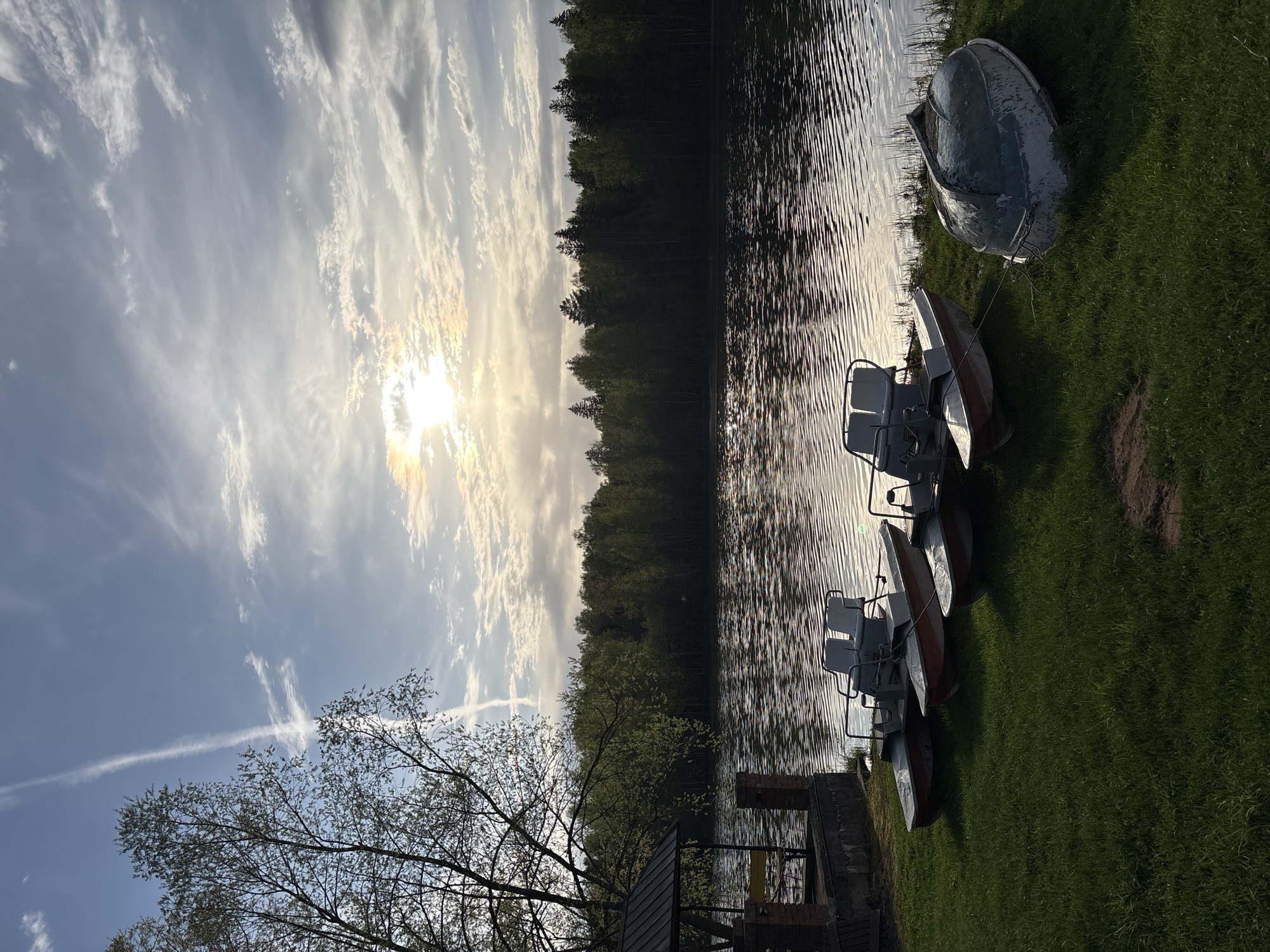 The image shows a view of a lake in Zavod Mikhailovskii, Russia. The sun is shining through a partly cloudy sky. The lake's surface reflects the sunlight and clouds. The edge of the lake is lined with trees and grass. Several pedal boats are parked on the grass beside the lake. One overturned metal boat is also on the grass. A small structure with a roof and brick pillars stands on the shoreline. A tree with sparse leaves is also in the foreground.