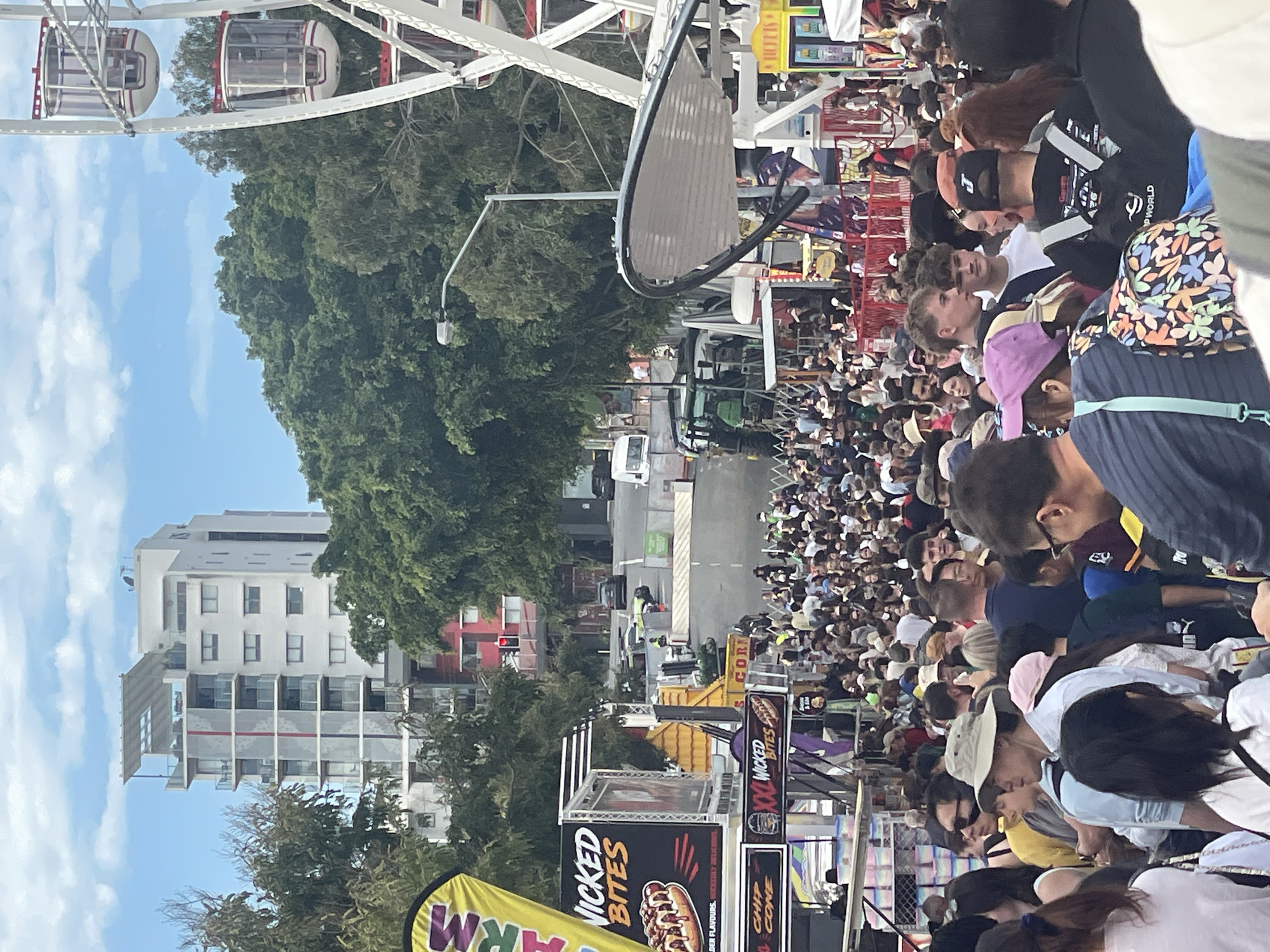 The image depicts a crowded outdoor event, likely a carnival or fair, in Bowen Hills, Australia, as indicated by its presence in the provided crops. In the background, a tall, modern residential building with balconies and a rooftop structure stands out against a blue sky with scattered clouds. Adjacent to the building, lush green trees form a dense canopy. To the right, a large Ferris wheel, partially visible, suggests an amusement park setting.
The foreground is dominated by a dense crowd of people of various ages and ethnicities, suggesting a popular event. They are standing and walking, some looking towards the camera. A variety of clothing is visible, including hats, t-shirts, and casual wear. Various stalls and temporary structures are set up, with signage indicating food vendors, including one advertising "WICKED BITES" with an image of a hot dog.
In the middle ground, a green tractor is positioned near some fencing and other fairground equipment. The scene appears to be taking place on a street or open area within the city. Traffic lights are visible in the distance, indicating a public road is nearby. The overall atmosphere suggests a lively and bustling event.