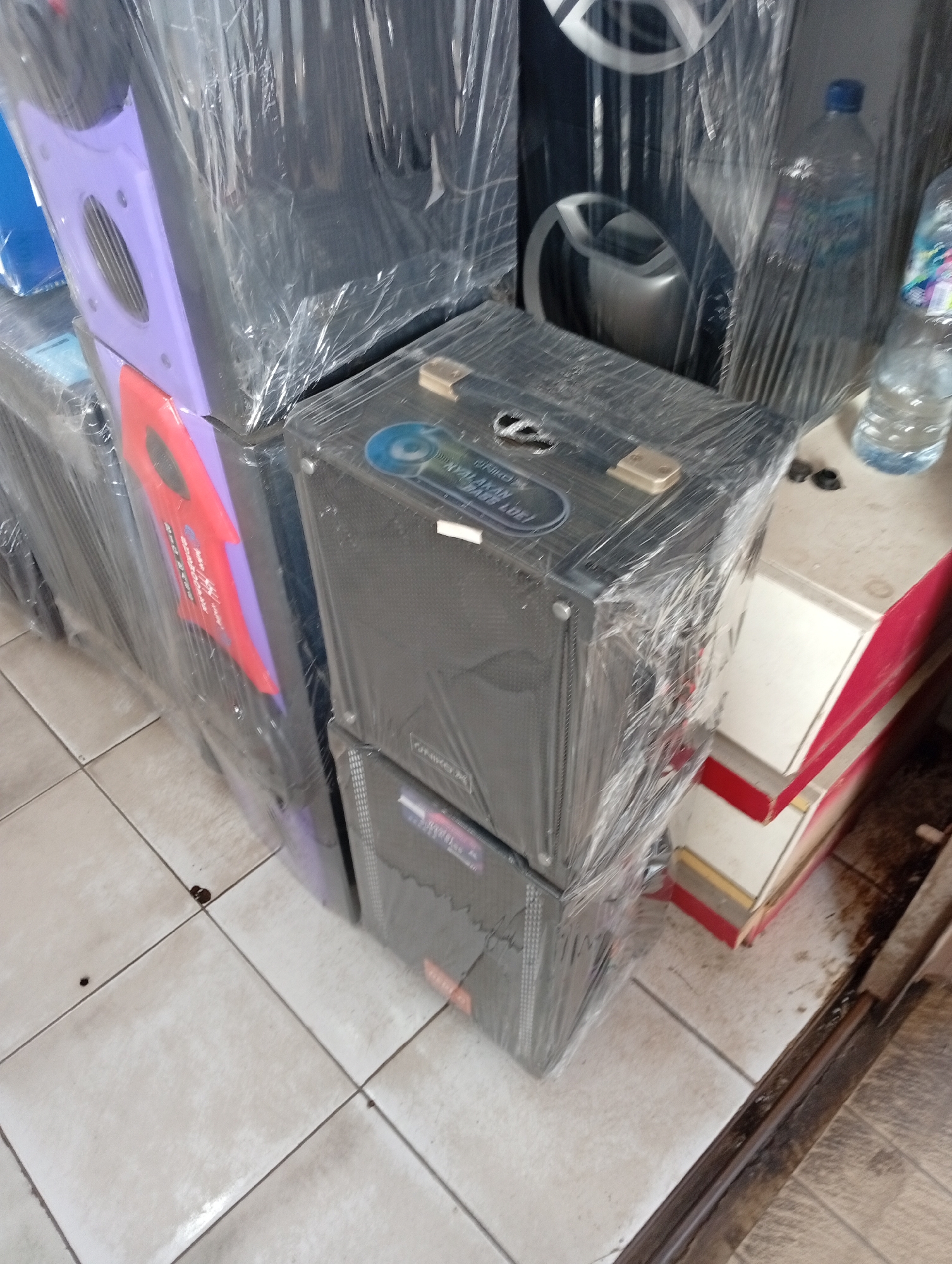 This image captures a close-up view of several stacked speakers, likely in a retail or storage environment. The speakers are wrapped in clear plastic, suggesting they are new or being protected. One speaker in the foreground, wrapped in plastic, has a prominent black grille and what appears to be a blue circular label with some text on it. To the left of this speaker, a larger, taller speaker unit with a purple section is visible, also wrapped in plastic. Further to the left, part of another large speaker with a red, guitar-shaped decorative element can be seen. In the background, another large speaker with silver accents is partially visible. Adjacent to the stacked speakers on the right, there are two clear plastic bottles of water and some other objects on a surface that appears to be a counter or shelf. The flooring beneath the speakers is tiled in a light color with visible grout lines. The overall impression is of an organized display or inventory of audio equipment. The image's context as being located in Batam, Indonesia, suggests a commercial setting within that city.