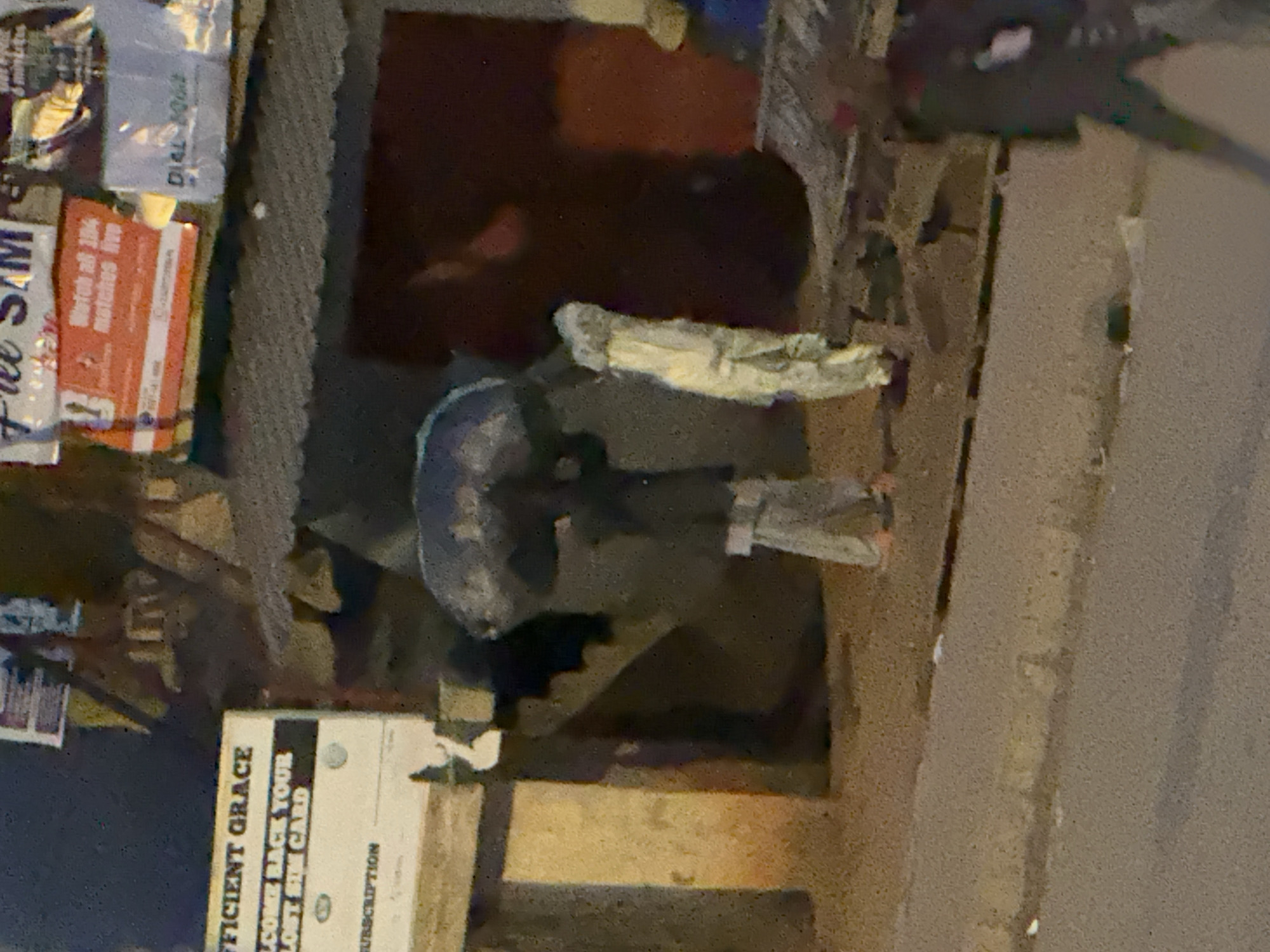 The image depicts a street scene in Bukuru, Nigeria, at night or under dim lighting. In the foreground, a blurred figure in dark clothing with white accents walks away from the camera. The midground shows two individuals. One person, holding a large, dark umbrella over their head, is dressed in a light-colored garment and appears to be walking. To their right, another figure, entirely covered in a white cloth, stands or walks. Behind them, a structure with a corrugated metal roof is visible, adorned with various posters and signs. One sign on the left reads "FFICIENT GRACE" and "WELCOME BACK YOUR LOST SIM CARD". Other signs are partially obscured or illegible. A rough, unpaved ground surface is present in the midground, transitioning to a paved road in the foreground. The overall impression is one of an urban environment experiencing some form of inclement weather, suggested by the umbrella.
