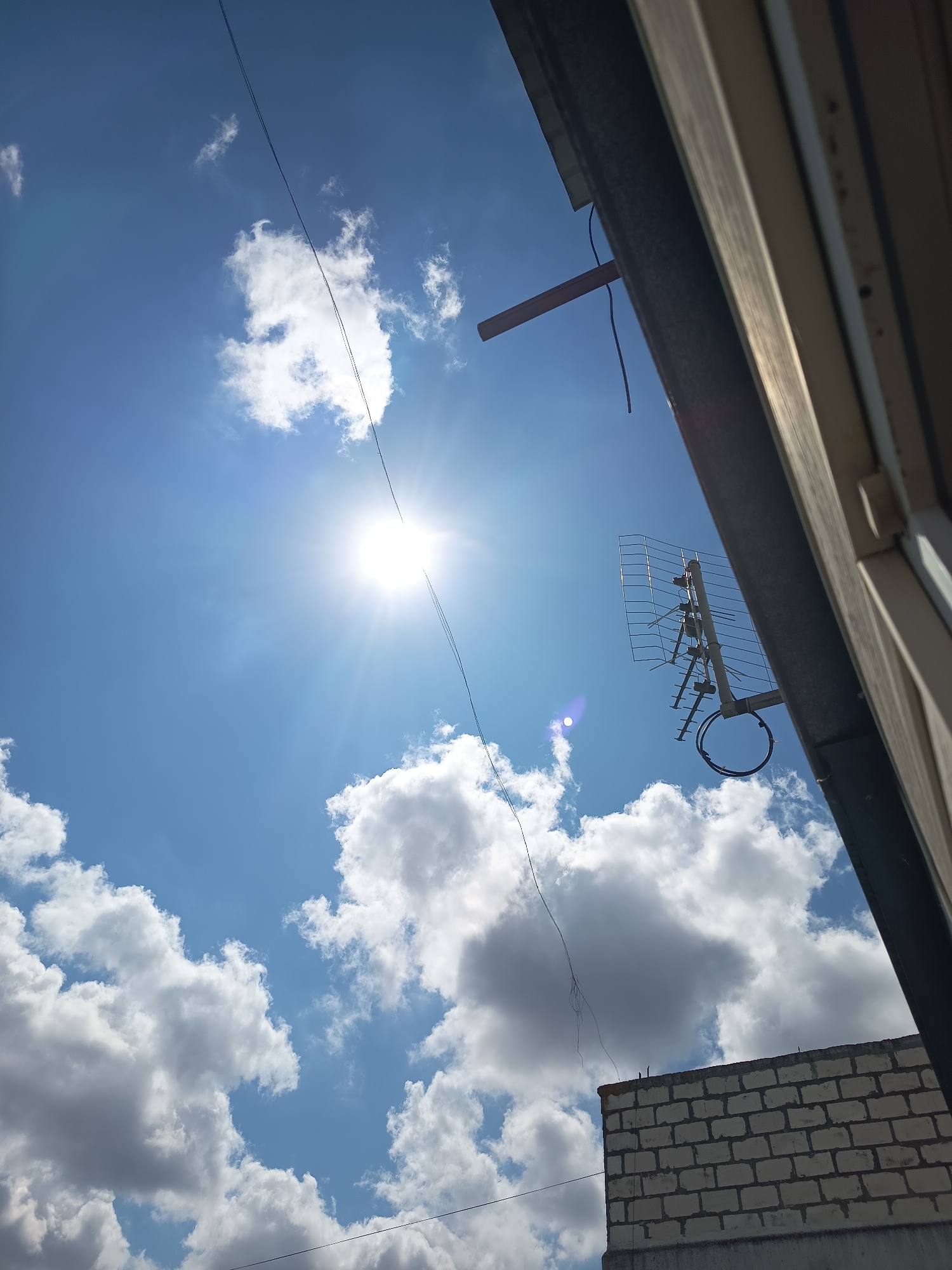 The image depicts a bright, sunny sky with scattered white clouds, viewed from a low angle. A prominent sun, with noticeable lens flare, shines in the upper left quadrant. A television antenna is mounted on a vertical pole to the right, with coiled cables at its base. A thin, dark wire runs diagonally across the sky from the upper left to the lower right. In the lower right corner, a portion of a brick wall is visible, suggesting an urban or suburban environment. The scene is captured outdoors, likely in the city of Izmail, Ukraine, given the provided context. No people or animals are present in the frame. The overall impression is a clear day with some man-made structures against a natural backdrop.