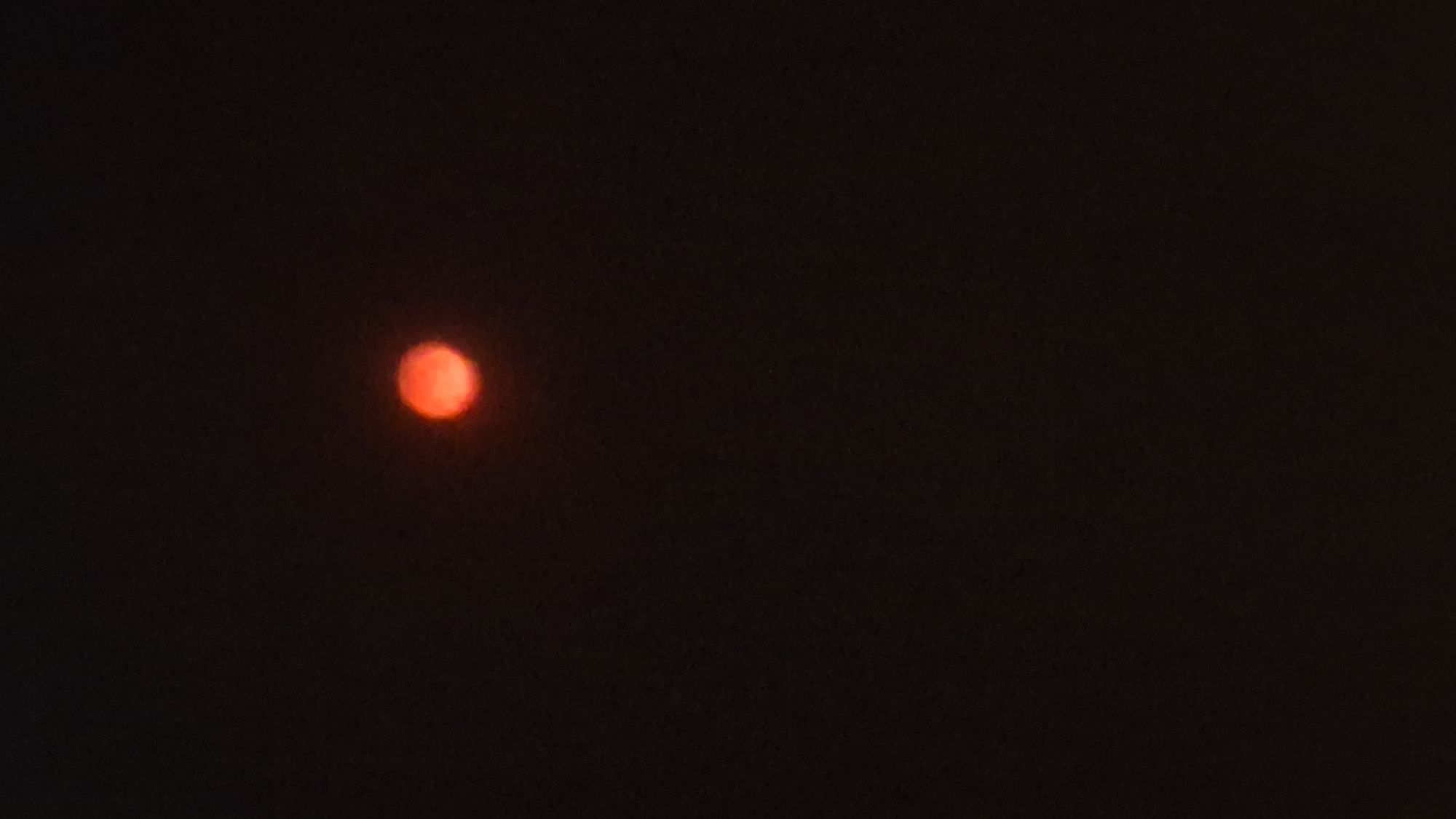 A solitary, fuzzy, reddish-orange orb is centered in a predominantly black, out-of-focus background. The orb, likely the sun filtered by atmospheric conditions such as smoke or dust, appears to be the only discernible object. Its indistinct edges and uniform hue suggest it is not a sharp celestial body but rather a diffused light source. The dark background is devoid of any discernible features or context, offering no clues about the environment or location. There are no people, animals, structures, or specific landmarks visible in the image. The overall impression is that of a celestial event observed through a heavily obscured atmosphere.