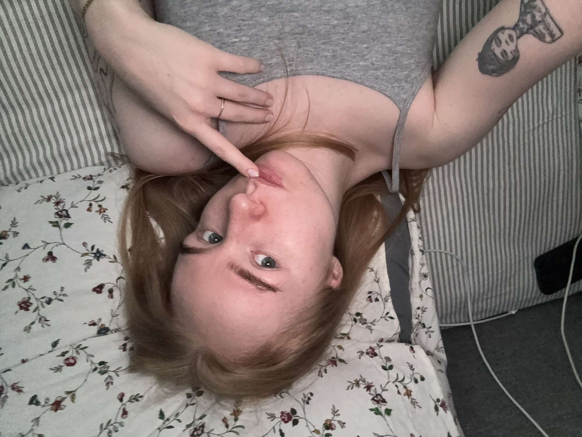 A fair-skinned, light-haired person with blue eyes lies on a bed. They are wearing a gray tank top. The person's left arm is raised, with a finger touching their lower lip. The right arm also is raised and the person has a tattoo on the upper arm. They are wearing a ring on their left ring finger. The bed has two pillow types, one is floral and the other is striped. There is a cable present next to a dark object. The photograph appears to be taken indoors with ambient lighting. The location is not identifiable as Chaikovsky, Russia.