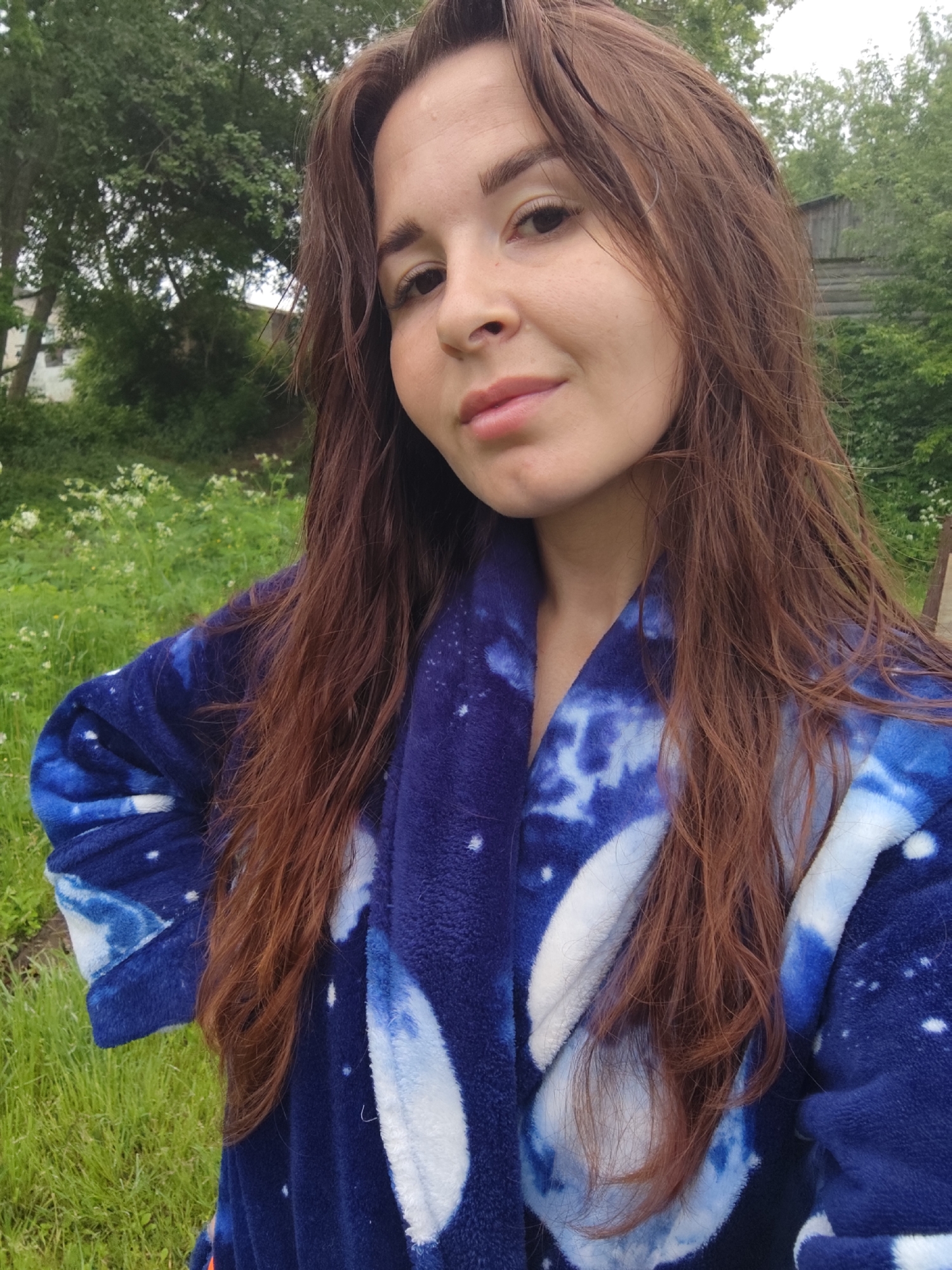 A young woman is featured in a close-up, likely a selfie, outdoors in what appears to be a rural or natural setting. She has long, wavy reddish-brown hair that frames her face and flows over her shoulders. Her dark eyes are directed towards the viewer, and she has a gentle, soft smile on her lips. She appears to be in her 20s or early 30s.
She is wearing a soft, plush robe or dressing gown. The robe is predominantly dark blue, adorned with intricate white and lighter blue patterns reminiscent of tie-dye, clouds, or celestial bodies like nebulae, along with scattered white speckles that resemble stars.
The background consists of vibrant green grass and dense foliage, including numerous trees, indicating an outdoor environment. Small white flowers are visible among the grass in the foreground. In the mid-ground, through the trees, a rustic wooden structure, possibly an old house or outbuilding, can be glimpsed, suggesting a traditional or natural landscape. The lighting is soft and diffused, indicating an overcast day or indirect daylight, possibly morning or late afternoon.
The woman's expression conveys a sense of calm and relaxation, enjoying the tranquil outdoor moment. The overall scene suggests a peaceful interlude in a natural or semi-rural area. The wooden structure and verdant environment align with the given location context of Фоки, Russia. There is no visible text in the image.