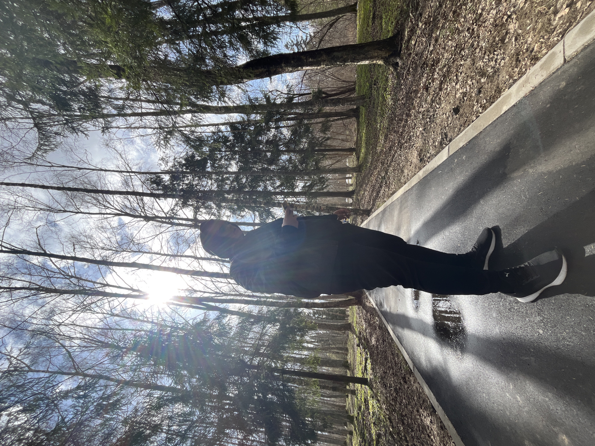 A person stands on a paved pathway in Vysokovsk, Russia, as evidenced by the surrounding landscape and architecture. The path is bordered by trees on one side and dead leaves on the other, suggesting an autumnal or early spring season. The person is wearing dark clothing and athletic shoes. A bright light source, likely the sun, is visible through the trees. There is a puddle on the path reflecting light.