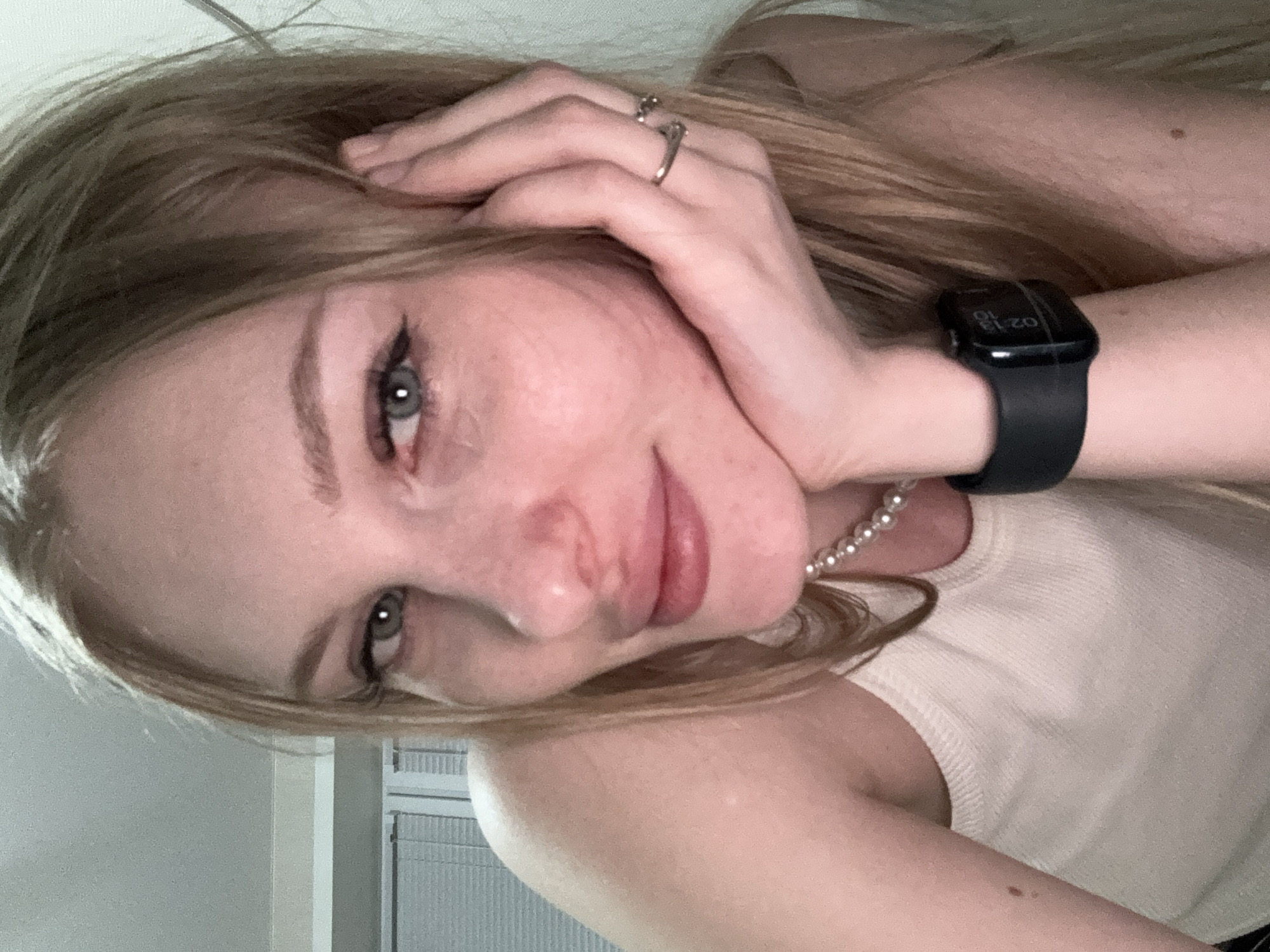 The image is a close-up, selfie-style shot of a young, fair-skinned woman with blonde hair. She is resting her chin on her hand, which is adorned with two rings. She is wearing a white tank top and a pearl necklace. A smartwatch is visible on her left wrist. The background is out of focus, but appears to be an indoor setting with blinds visible on the left. There are no specific landmarks or indicators within the image that would allow for the determination of a location in Perm, Russia. The lighting suggests an indoor environment, possibly at night or in low light conditions.
