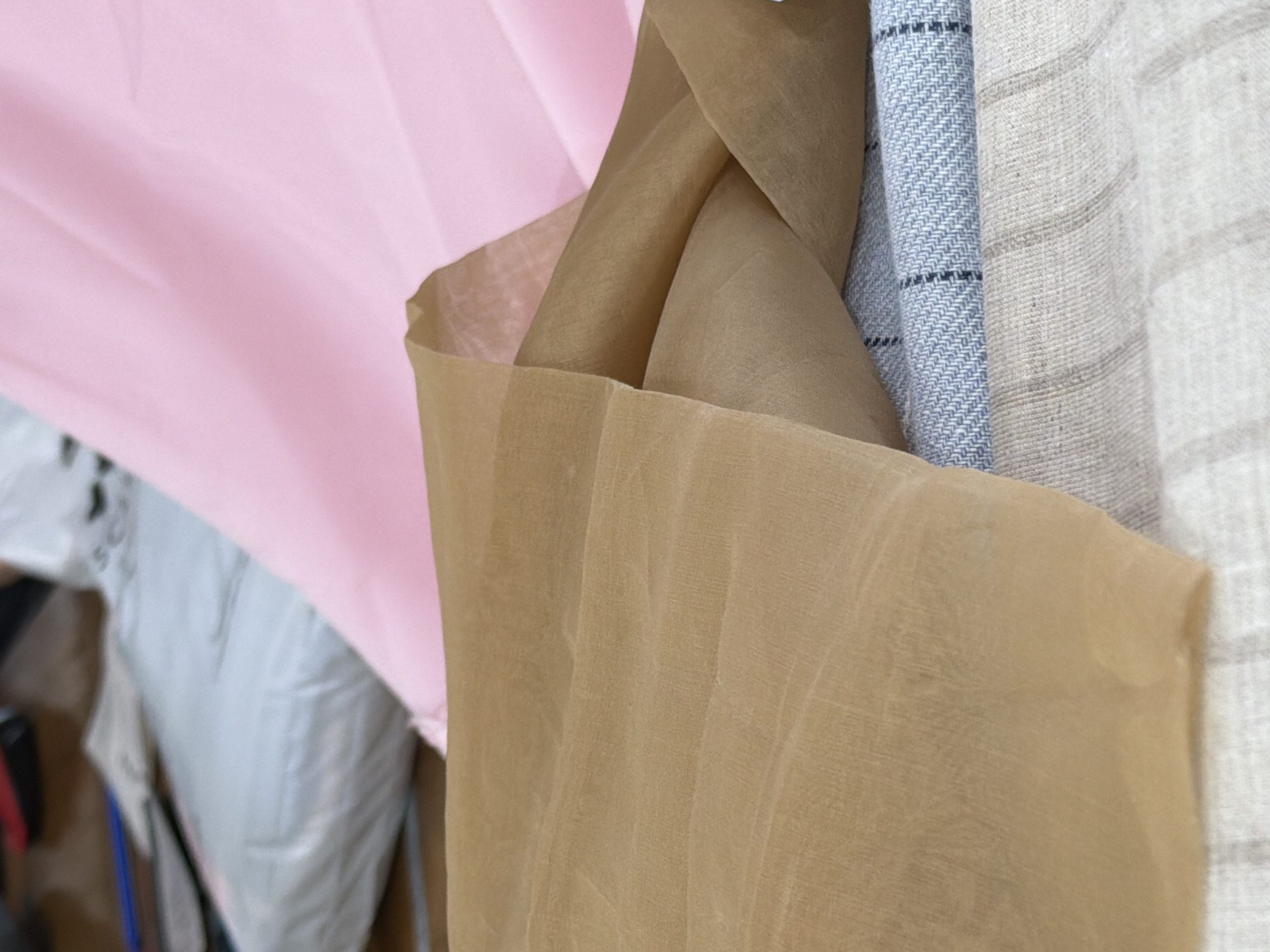 The image displays a close-up, elevated view of multiple textile swatches and bundled fabrics arranged indoors, in Saint Petersburg, Russia. In the immediate foreground and mid-ground, a large piece of translucent, sheer tan-colored fabric is prominently draped, exhibiting subtle wrinkles and folds. Beneath this, a stack of various patterned and textured fabrics is visible. The top fabric in the stack is light blue with a herringbone weave and horizontal dark blue stripes. Below it, a light beige or natural-toned fabric with subtle vertical striped patterns is present, followed by another lighter-toned, possibly off-white, striped fabric at the bottom. In the upper right background, a section of soft pink fabric is draped, creating a gentle contrast. To the upper left, blurred background elements include a light grey or white packaged item, possibly another textile bundle, along with indistinct darker objects, suggesting a cluttered work surface or storage area. No individuals are visible. The lighting is soft and diffused, highlighting the textures of the textiles.