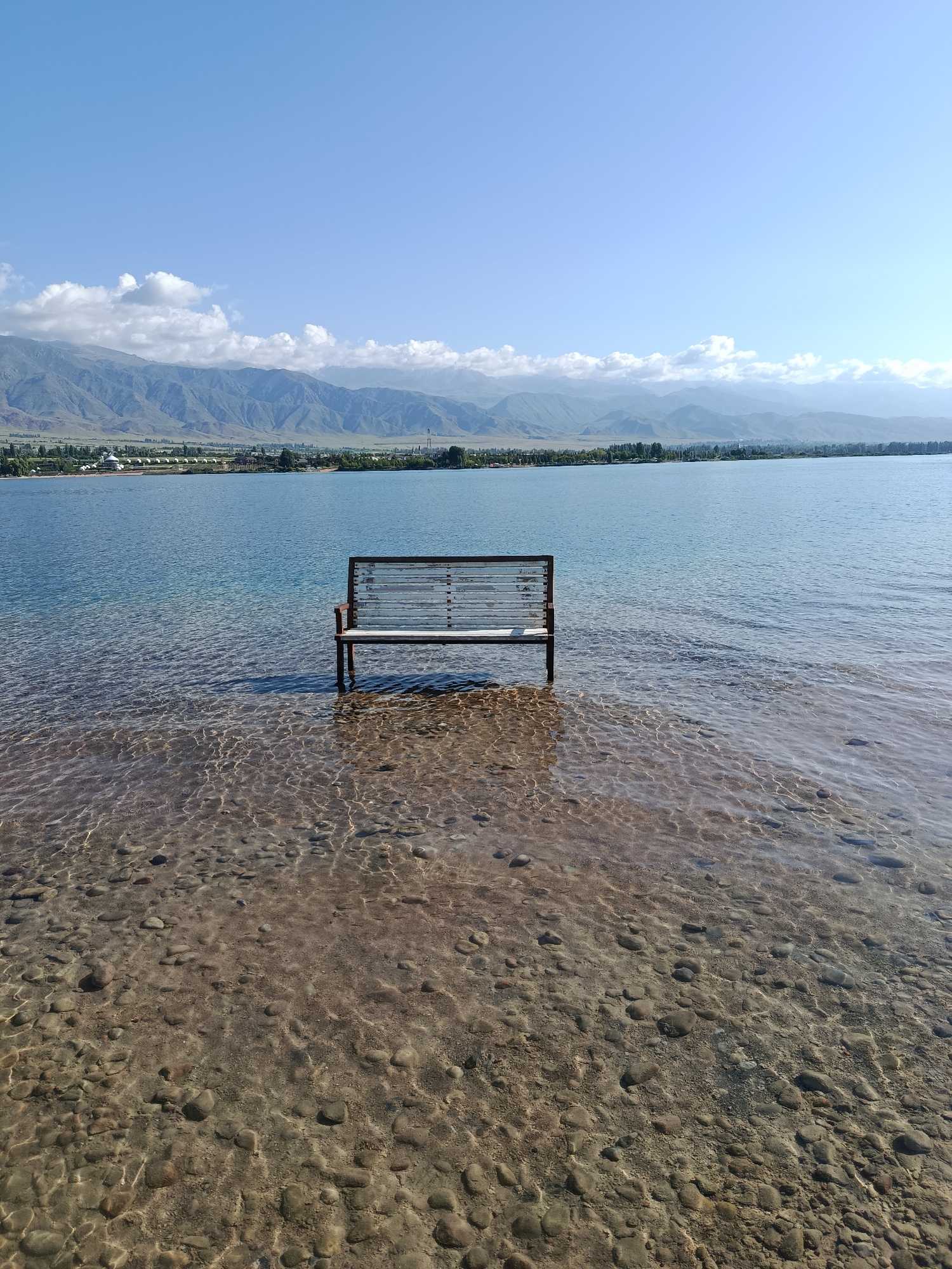 An empty, wooden bench sits partially submerged in the clear, shallow water of a lake, with the rocky lakebed visible beneath the surface. The water stretches to a distant shoreline, where trees and buildings are visible against a backdrop of hazy mountains under a bright blue sky with scattered clouds. The scene is serene and still.