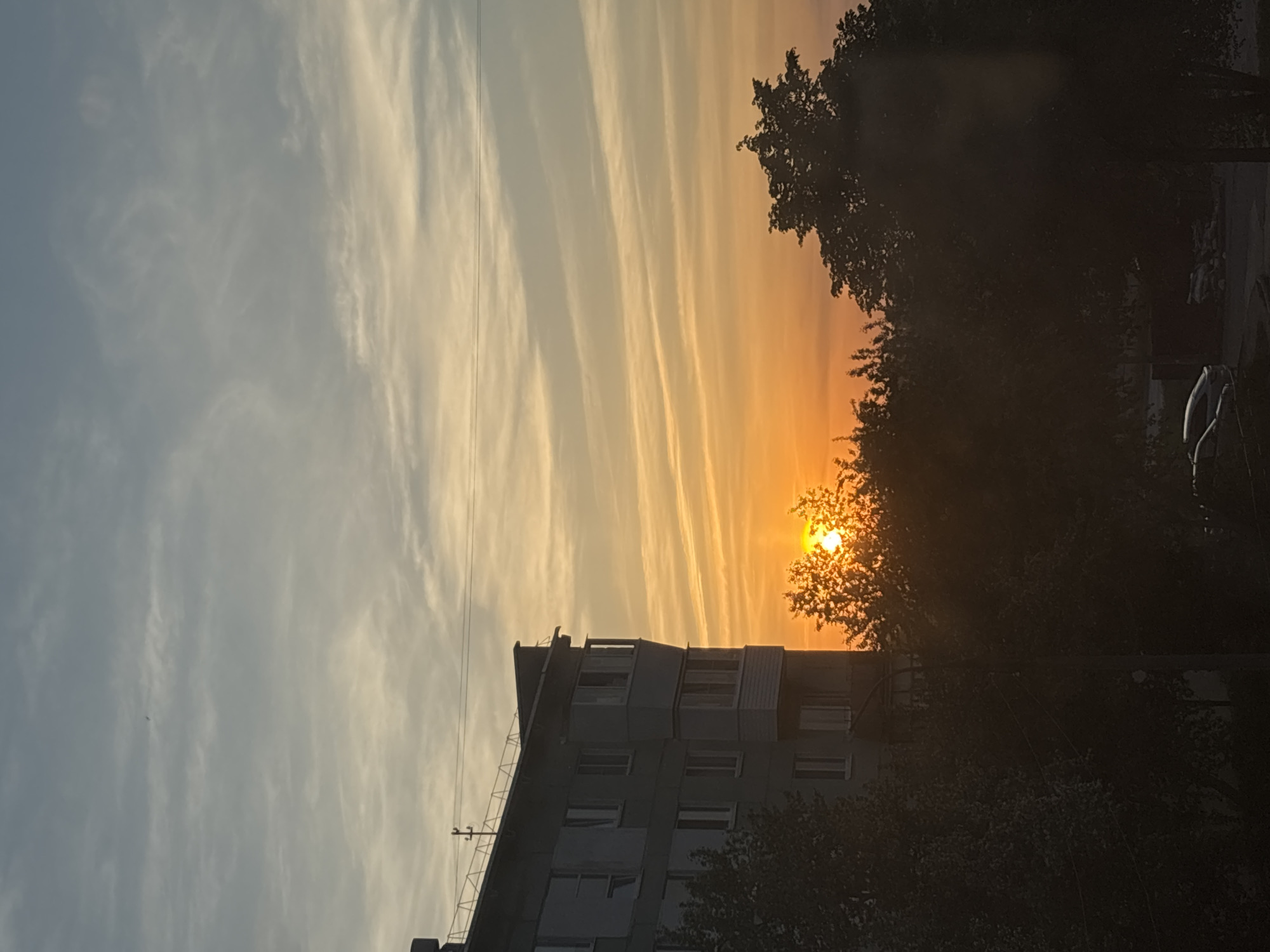 The image depicts a sunset in what is suspected to be the city of Chaikovsky, Russia. The sun is positioned behind a tree, casting a warm orange glow in the sky. The sky has striped clouds. A building is visible in the foreground. Vegetation and a car are present on the right. The image is tilted and appears to have been taken at ground level.