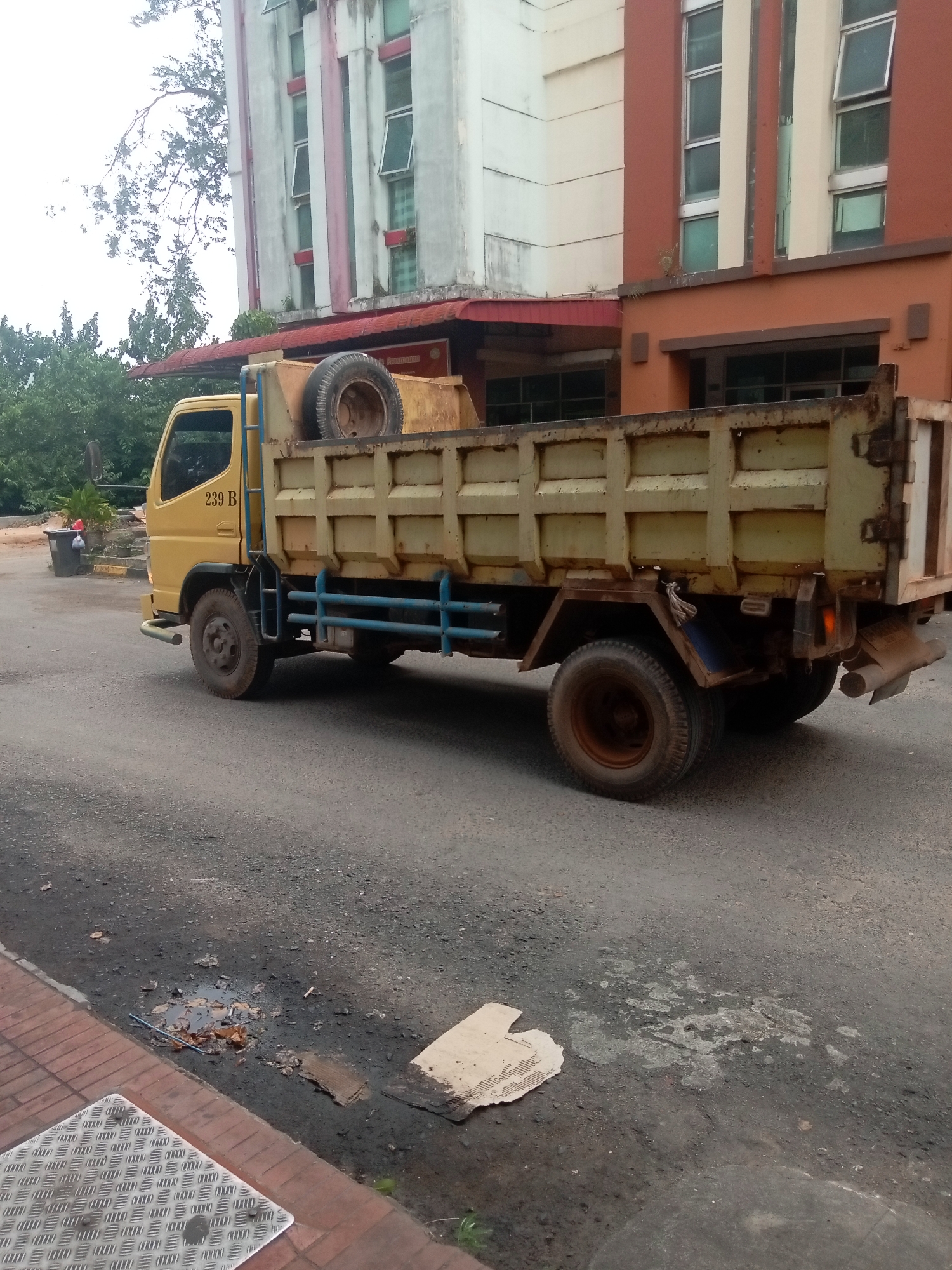 A yellow dump truck with a worn bed is parked on an asphalt surface. The truck has a spare tire mounted in the front of its bed and blue metal railings along the sides. A white building with brown accents and multiple windows is visible behind the truck. The ground in front of the truck is paved with bricks and has a metal grate. There are some puddles and debris on the asphalt. The scene appears to be in an urban area of Batam, Indonesia.