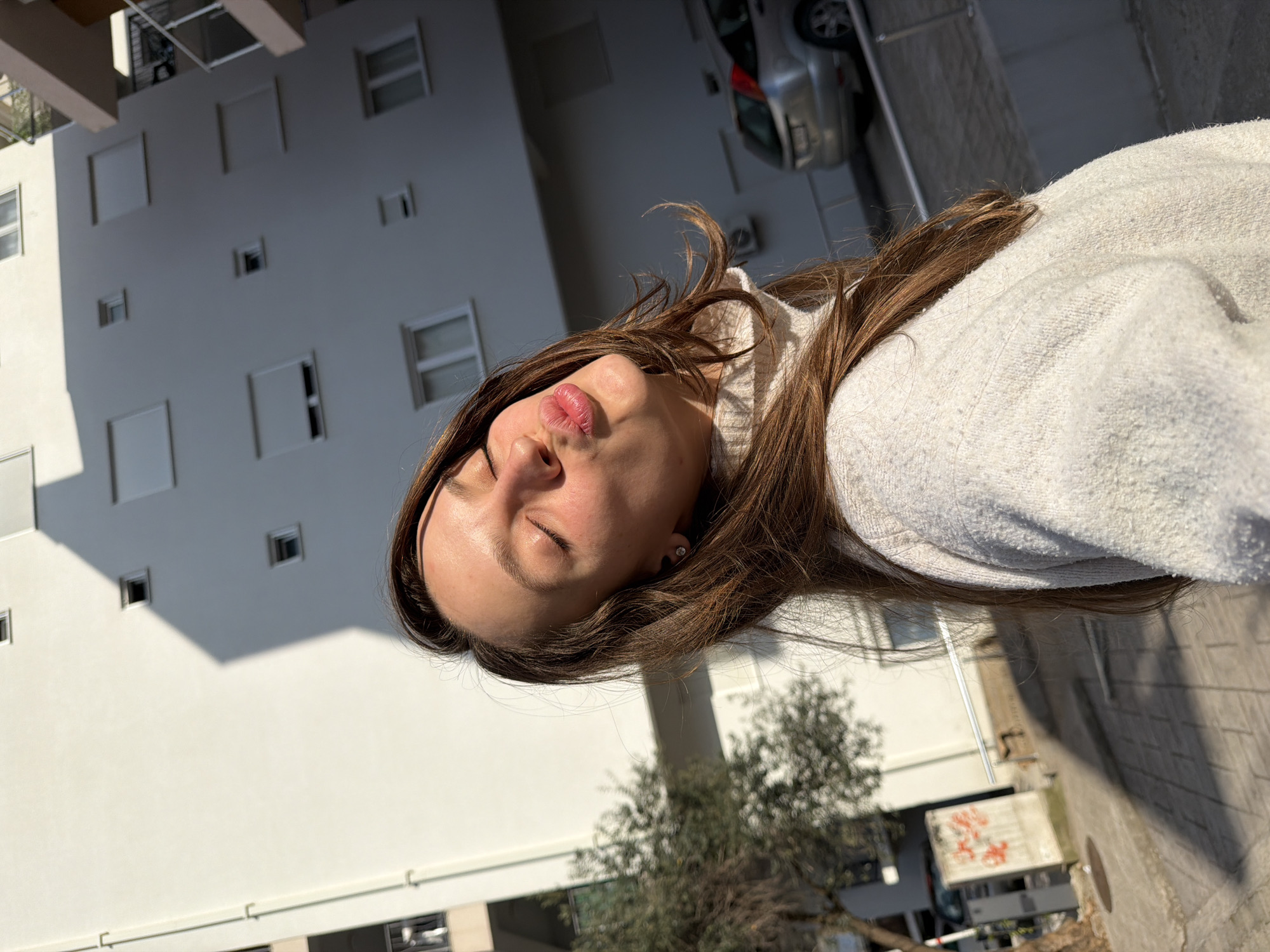 A medium shot, taken outdoors in daylight, shows a young woman in a white hoodie with closed eyes and pursed lips, positioned in the foreground. Behind her, a tall, white building with multiple small windows and a partial view of a silver car parked on a paved street. A small tree is visible to the left of the building. The location is likely Budva, Montenegro, given the architectural style of the building and the Mediterranean vegetation. The shot appears to have been taken at an unusual angle, possibly from a lower vantage point, looking upward.