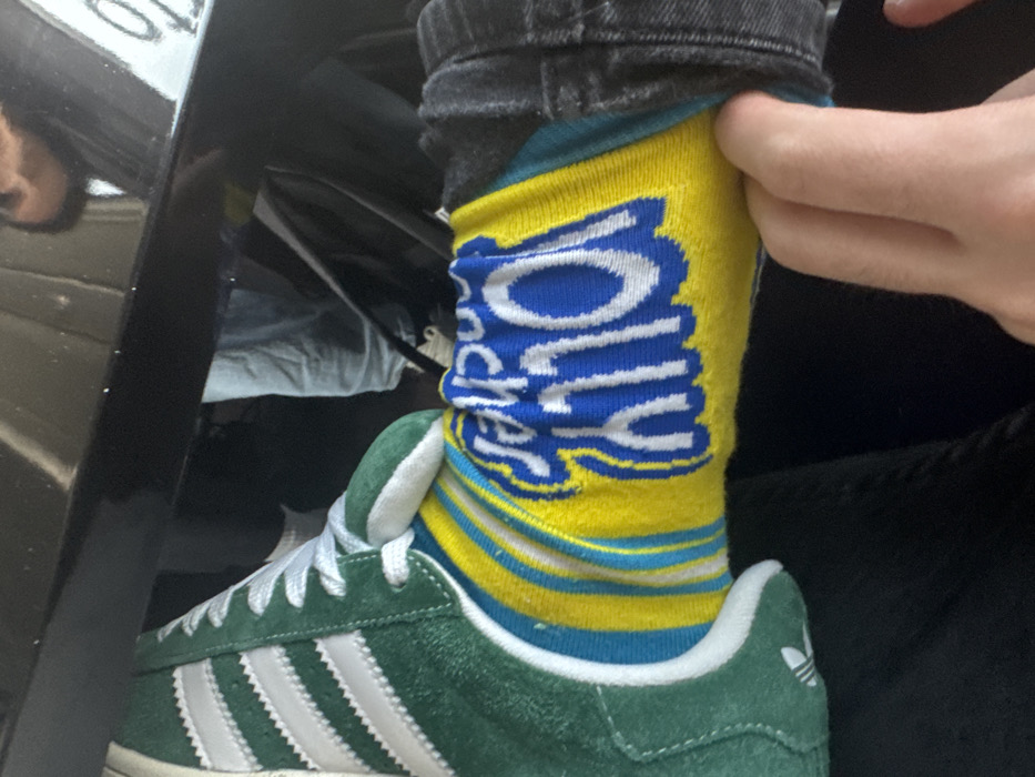 A person wears green suede Adidas sneakers, with white stripes and laces, paired with yellow and blue “Jolly Rancher” branded socks. Their jeans are dark gray. A hand pulls at the top of the sock, adjusting it. Part of a dark, possibly black, car panel is visible to the left. The photo appears to be taken indoors.